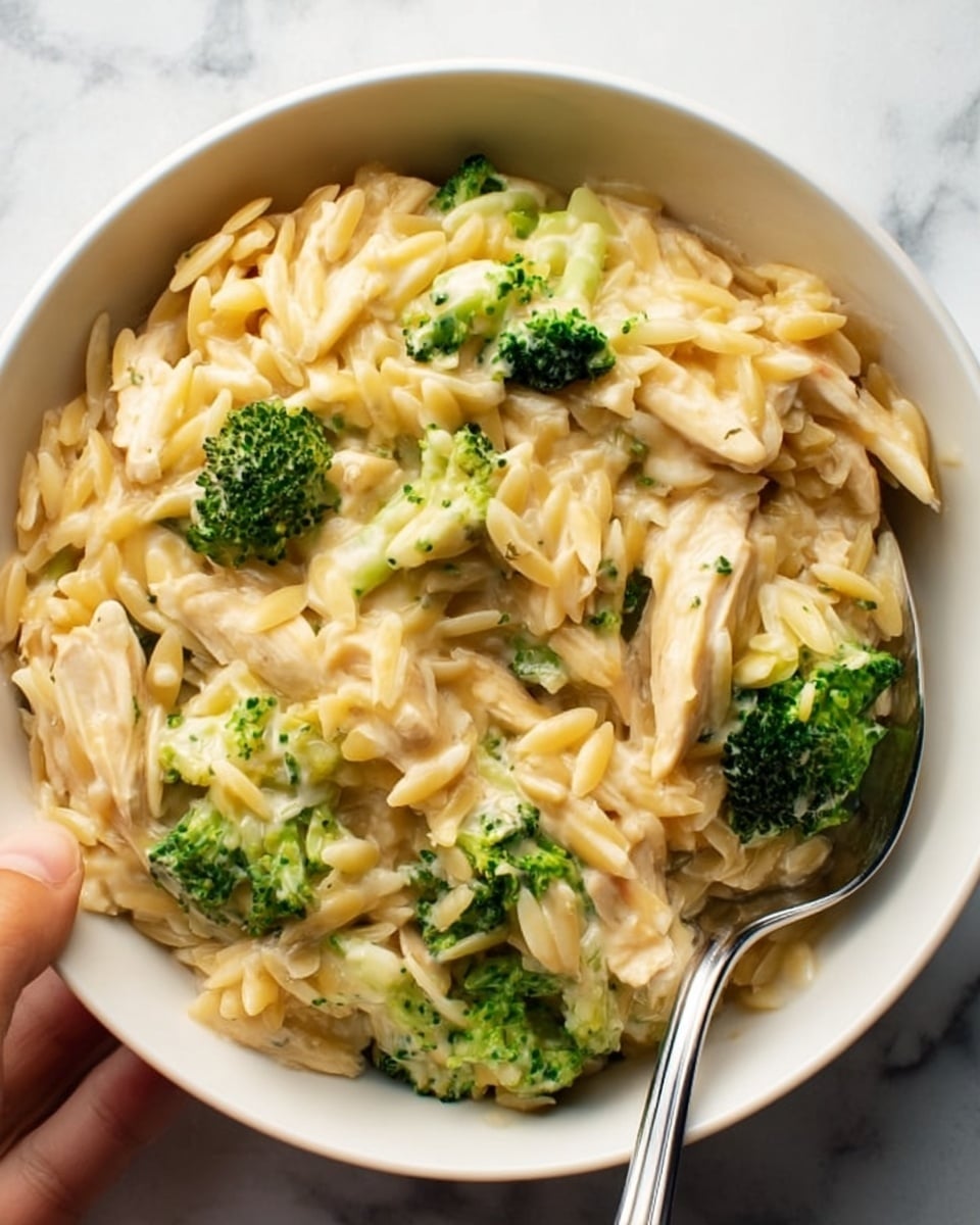 This image shows a white bowl filled with creamy pasta mixed with shredded light-colored chicken and small bright green broccoli florets. The pasta is small and looks smooth with a pale yellow sauce coating every piece, blending well with the tender chicken strips and green broccoli. A shiny silver spoon rests in the bowl, partially covered by the creamy mix, and a woman's hand holds the handle. The background surface is a white marbled texture, adding a clean and fresh look to the scene. photo taken with an iphone --ar 4:5 --v 7
