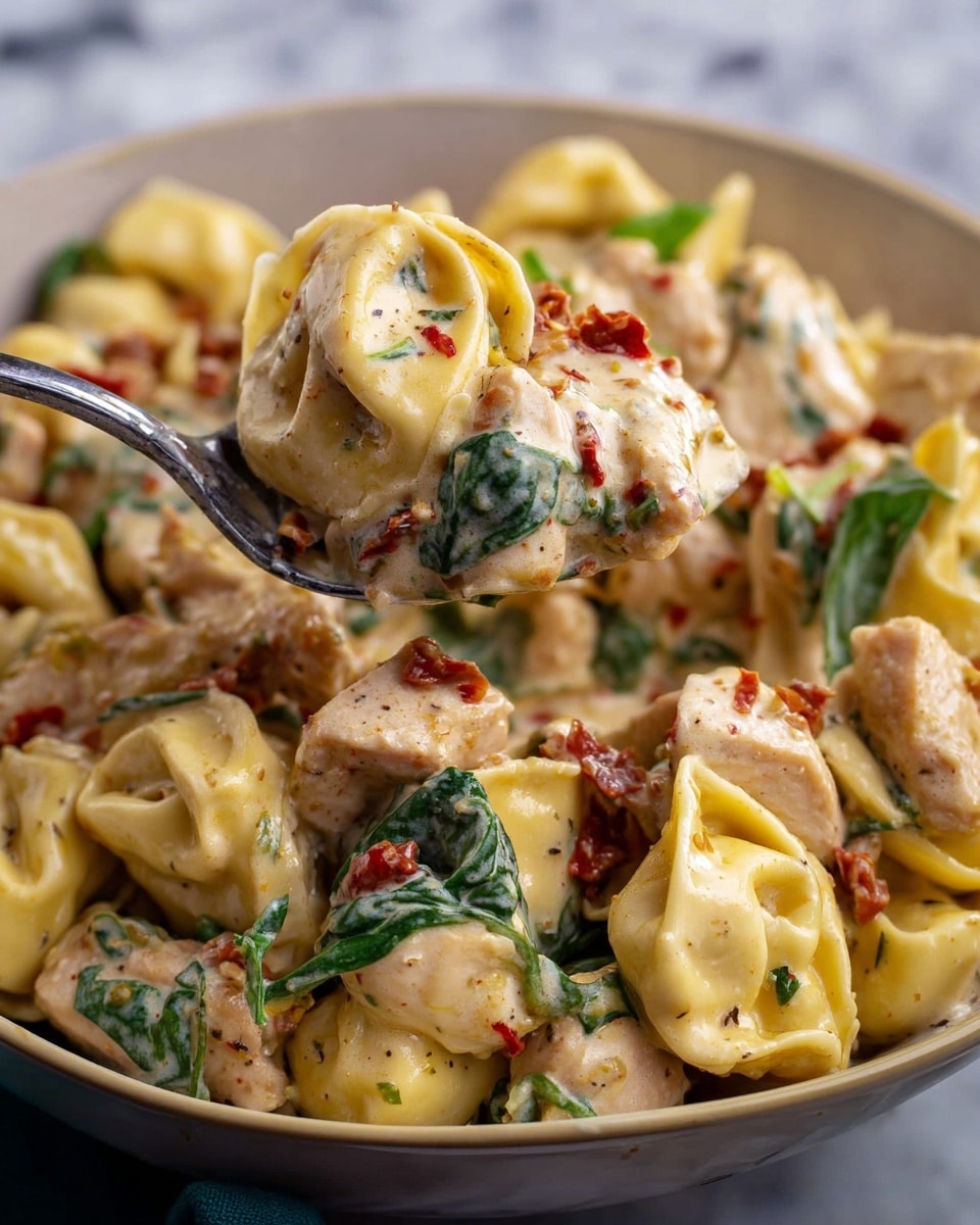 The image shows a close-up of a spoon lifting one layer of yellow tortellini pasta covered in creamy white sauce with small red bits and pieces, mixed with chunks of light brown cooked chicken and fresh green spinach leaves scattered throughout. The tortellini are plump and smooth with a slightly shiny texture from the sauce. The dish is served in a white bowl placed on a white marbled surface, highlighting the warm and rich colors of the pasta and sauce. Photo taken with an iphone --ar 4:5 --v 7