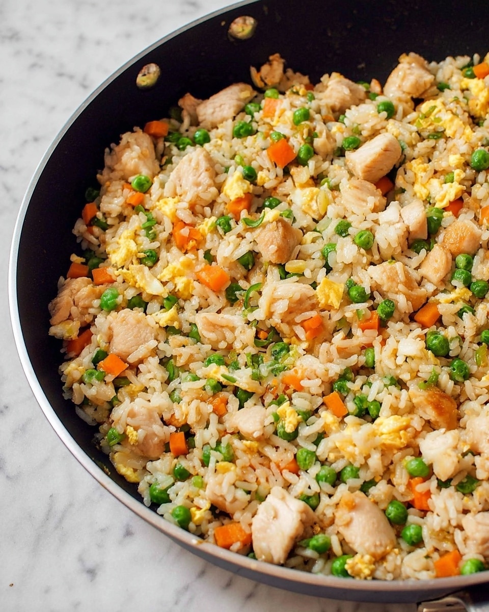 A close-up view of a large black frying pan filled with fried rice, showing layers of white rice mixed with small green peas, orange carrot cubes, light tan pieces of cooked chicken, and soft scrambled egg bits in light yellow. The fried rice looks well mixed, with a slightly shiny texture on the surface. The pan is placed on a white marbled surface. photo taken with an iphone --ar 4:5 --v 7