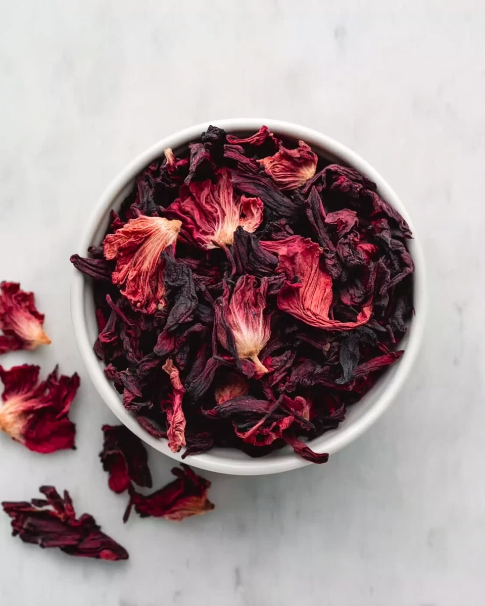 A white bowl filled with dried hibiscus flowers in dark red and deep purple shades, each petal having a crinkled texture. Some dried petals lie scattered beside the bowl on a white marbled surface, adding a natural and slightly messy look. The image is viewed from above, showing the mix of colors and textures clearly. Photo taken with an iphone --ar 4:5 --v 7