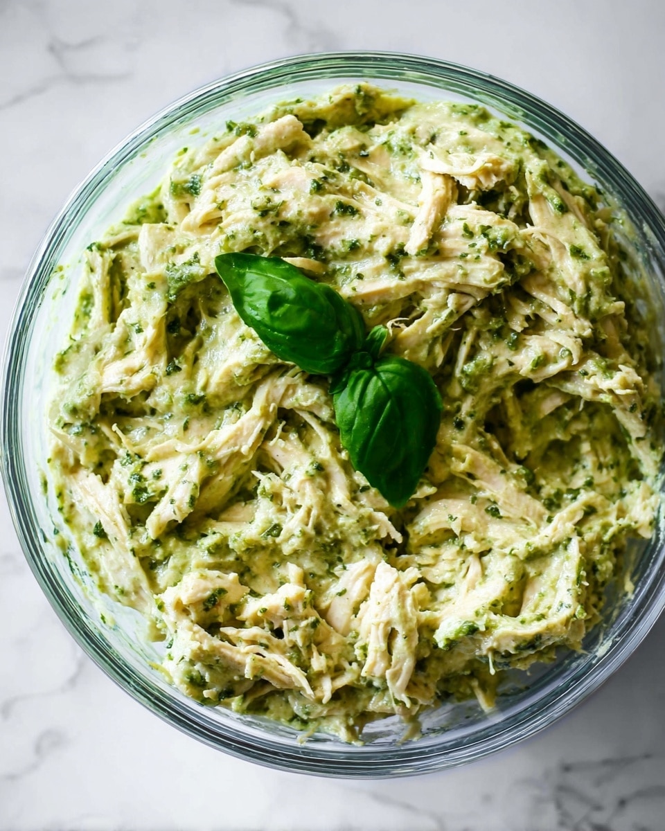 A clear glass bowl filled with a creamy mix of shredded white chicken and green pesto sauce, creating a thick, textured appearance with visible strands of chicken coated in a smooth green mixture. The top is garnished with two fresh basil leaves, adding a bright dark green contrast. The bowl is placed on a white marbled surface. photo taken with an iphone --ar 4:5 --v 7