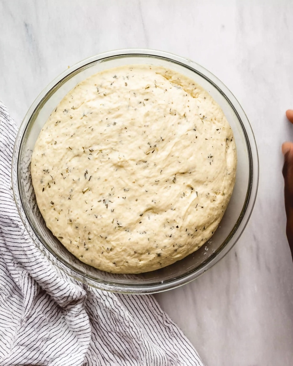 The image shows a round glass bowl filled with dough that has risen and doubled in size. The dough is light creamy color with small black herb specks scattered throughout. The dough has a soft, smooth, and slightly bumpy texture on the top surface, stretching evenly inside the bowl. The glass bowl sits on a white marbled surface next to part of a white and gray-striped cloth. In the top right corner, you can see a woman’s hand gently touching or near the bowl. Photo taken with an iphone --ar 4:5 --v 7