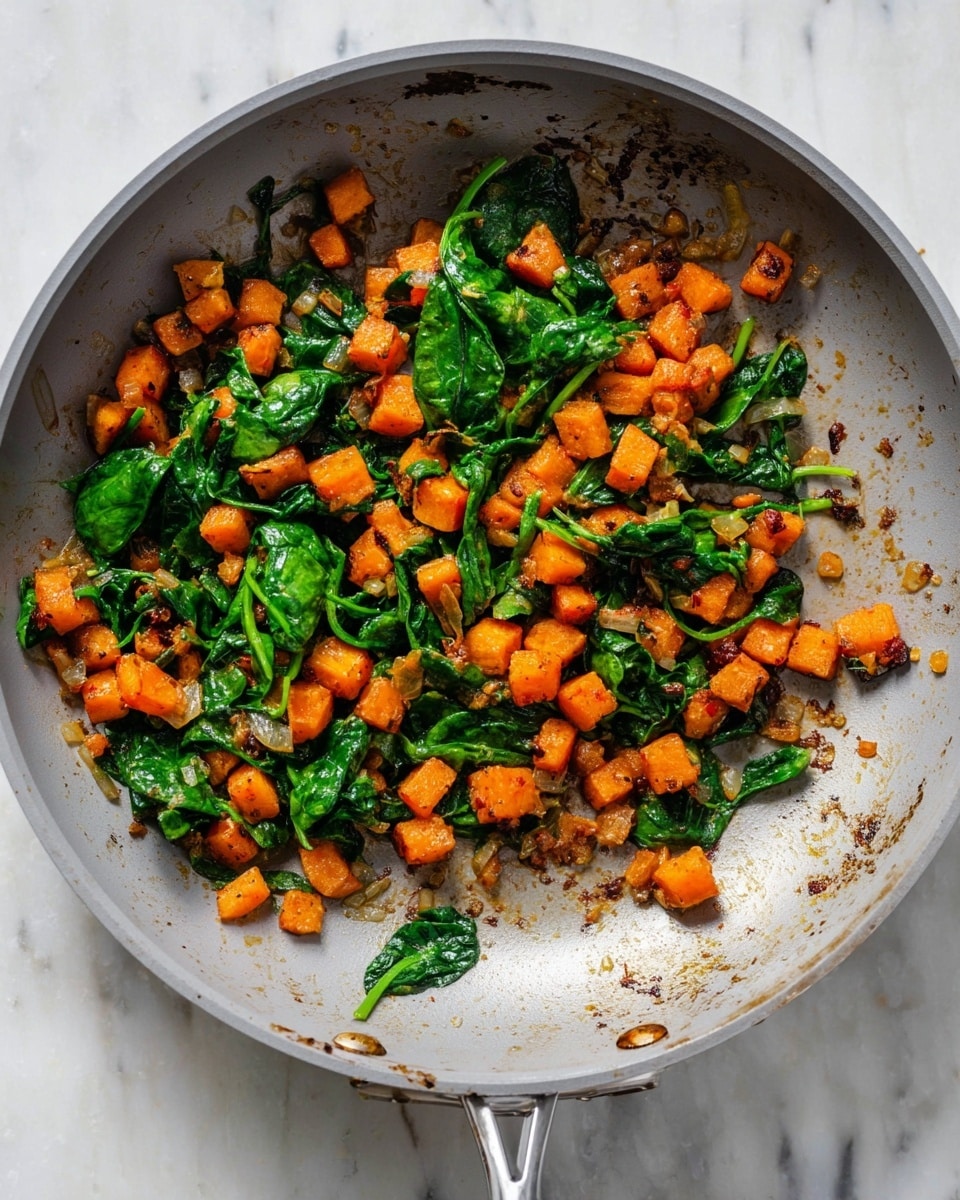 A close-up view of a frying pan with a cooked mix of small orange cubes and fresh green spinach leaves inside. The orange cubes are soft and lightly browned, scattered evenly throughout the pan. The spinach leaves are wilted but still hold their deep green color, mixed naturally with the orange cubes. Small bits of translucent cooked onion are visible among the vegetables. The pan has a smooth gray interior and a shiny metal handle with some cooked food marks inside. The pan sits on a white marbled surface, enhancing the colors of the vegetables. photo taken with an iphone --ar 4:5 --v 7