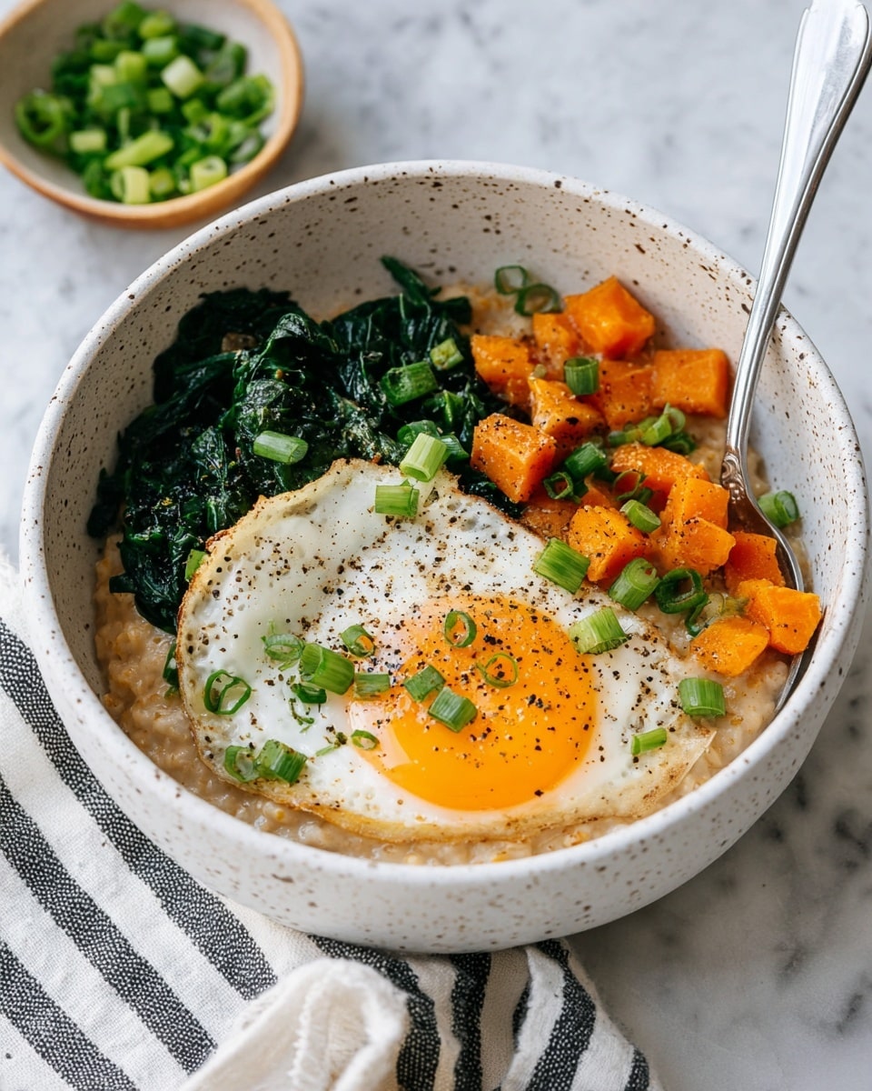 A white speckled bowl holds a layered dish starting with a creamy, light brown base of oatmeal filling the bottom half, topped on the left by a sunny-side-up egg with a bright orange yolk and white edges sprinkled with black pepper and chopped green onions. To the right, sautéed dark green spinach and small orange cubes of cooked sweet potatoes sit side by side, adding bold color contrast. A silver spoon is placed inside the bowl near the egg, and a small bowl of chopped green onions is visible in the background on a white marbled surface with a striped cloth nearby. Photo taken with an iphone --ar 4:5 --v 7