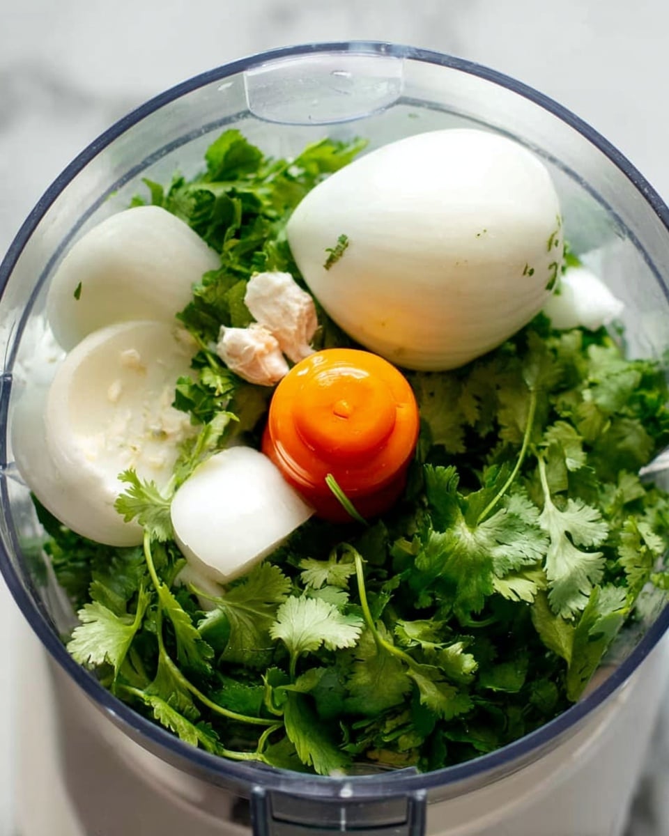 Inside a clear food processor bowl, there are four layers of ingredients. At the bottom are bright green fresh cilantro leaves with visible detailed textures and thin stems. On top of the cilantro, there are several white onion pieces with smooth, round, curved surfaces. Nestled among the onion pieces are two small white garlic cloves with tiny brown tips. At the top center, there is one small, bright orange pepper with a slightly wrinkled texture. The background surface is white marble. photo taken with an iphone --ar 4:5 --v 7