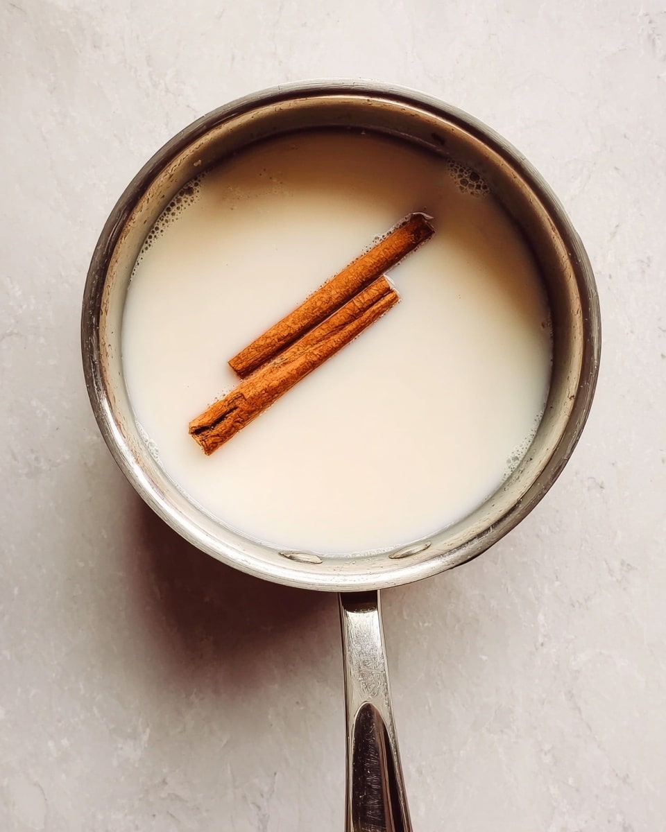 A shiny silver saucepan sits on a white marbled surface, filled with a creamy white liquid. Two brown cinnamon sticks float gently on top, adding contrast to the smooth liquid. The saucepan handle points downwards, and the image is lit softly to show the creamy texture and the wooden detail of the cinnamon sticks. Photo taken with an iphone --ar 4:5 --v 7