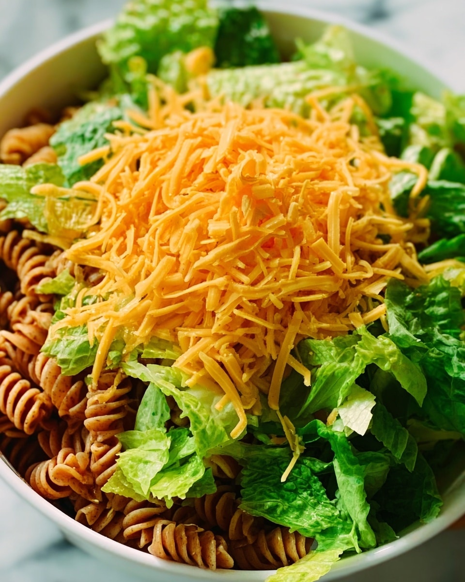 The image shows a close-up of a white bowl filled with a layered salad. The bottom layer has cooked spiral pasta with a brownish color, spread evenly at the base of the bowl. On top of the pasta, there are fresh green lettuce leaves arranged loosely and covering most of the pasta. The top layer is a large pile of finely shredded orange cheese placed in the center, slightly spilling over the lettuce. The bowl is placed on a white marbled surface. Photo taken with an iphone --ar 4:5 --v 7