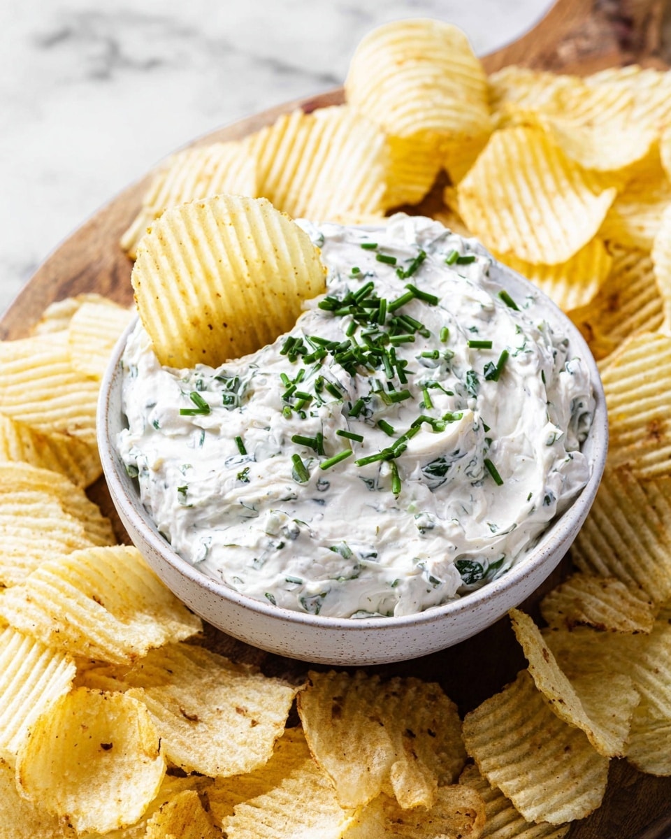 A white bowl filled with a creamy, thick dip speckled with small green herb pieces, topped with chopped fresh green chives. A ridged potato chip is dipped halfway into the creamy dip, showing its smooth texture. The bowl sits on a wooden board surrounded by many ridged and plain light yellow potato chips. The entire scene is on a white marbled surface. photo taken with an iphone --ar 4:5 --v 7