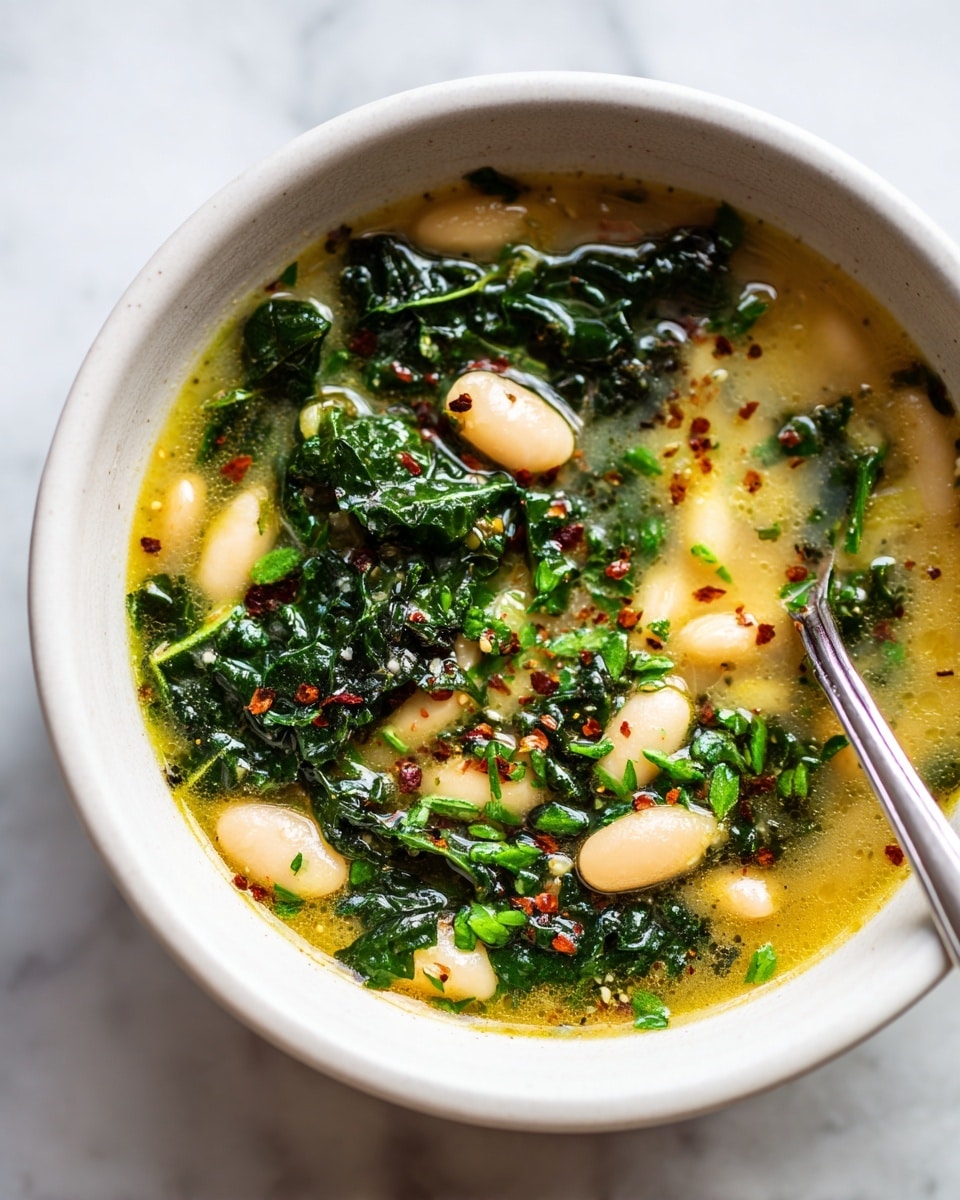 A close-up view inside a white bowl filled with a thick soup showing three main layers: a pale yellow broth base with a smooth texture, scattered white beans floating throughout, and fresh, dark green chopped kale leaves spread evenly on top. Small green herb pieces are sprinkled across the surface with fine red spice flakes adding specks of color. A silver spoon rests inside the bowl partly submerged, catching light and reflecting the ingredients. The background is a white marbled texture. Photo taken with an iphone --ar 4:5 --v 7