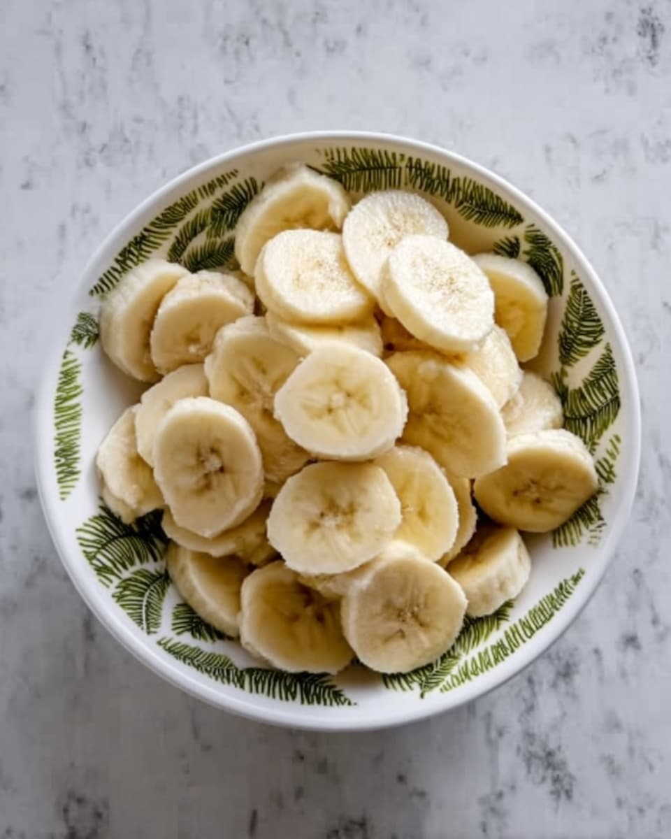 A white bowl filled with many slices of banana, showing light yellow and creamy textures. The bowl has a simple green leaf pattern on the edge. The bananas look fresh and soft, layered over each other, creating a rounded, full look in the bowl. The bowl is placed on a white marbled surface with subtle gray veining. Photo taken with an iphone --ar 4:5 --v 7