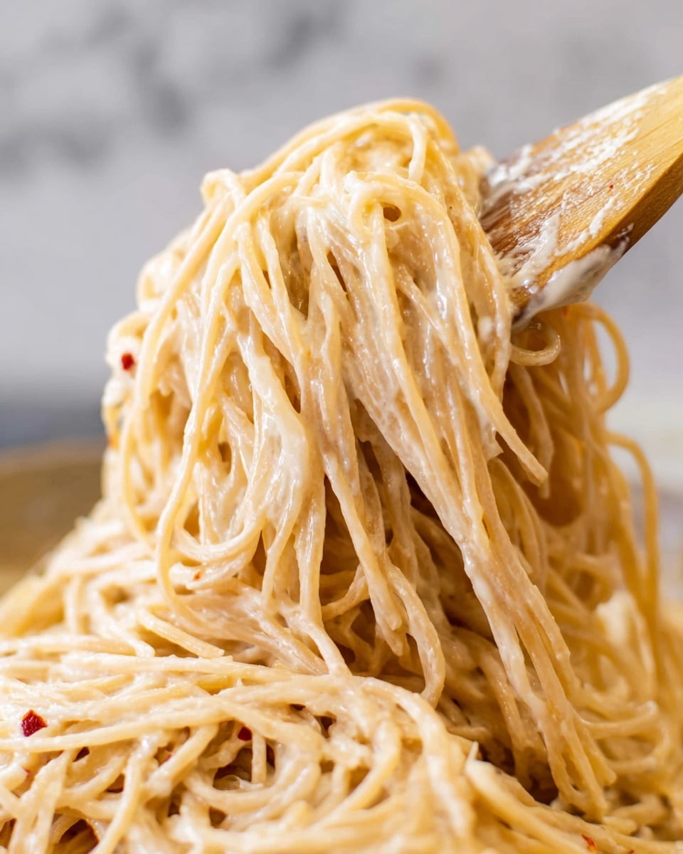 A close-up view of a large clump of spaghetti noodles coated in a creamy, light beige sauce, lifted by a wooden spatula covered lightly with the sauce. The spaghetti strands are soft and tangled, showing some small red specks, possibly chili flakes, scattered throughout. The background is softly blurred, highlighting the noodles' smooth and glossy texture and the wooden spatula’s worn surface. The scene sits on a white marbled texture. photo taken with an iphone --ar 4:5 --v 7