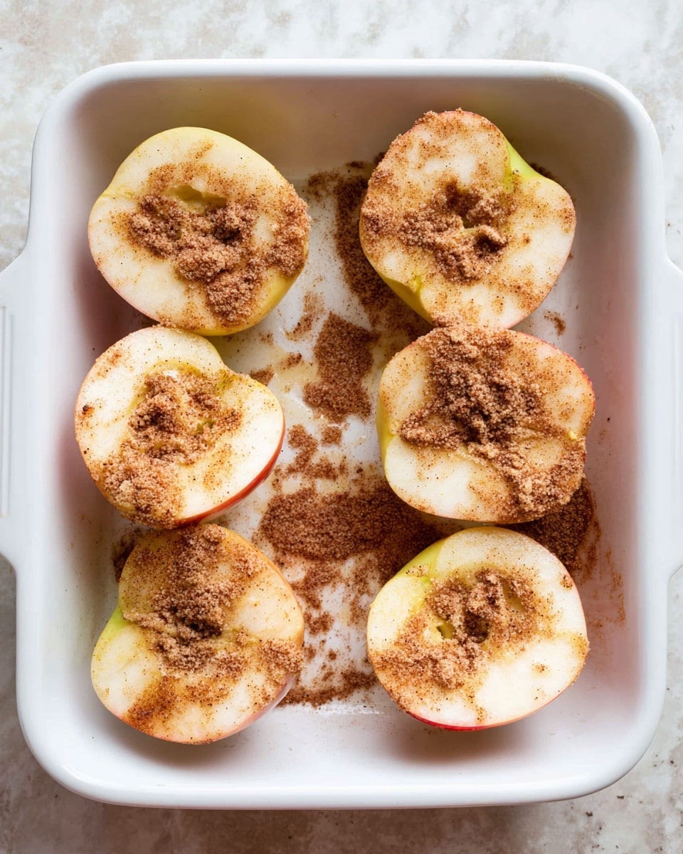 A white square baking dish holds six apple halves arranged in two rows of three, each apple half showing its pale cream inside with touches of red on the edges. The apple halves are sprinkled generously with a light brown, slightly clumpy cinnamon and sugar mixture that covers their inner surfaces unevenly, creating a textured, powdery effect. The dish sits on a surface with a white marbled texture. photo taken with an iphone --ar 4:5 --v 7