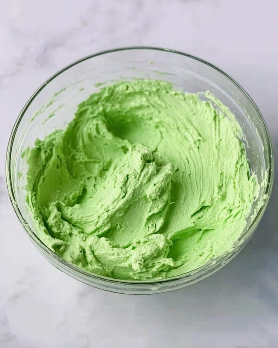 A clear glass bowl filled with a thick, bright green creamy mixture that has a smooth, slightly swirled texture. The green color is even throughout, and the mixture appears fluffy and soft. The bowl is placed on a white marbled surface. photo taken with an iphone --ar 4:5 --v 7