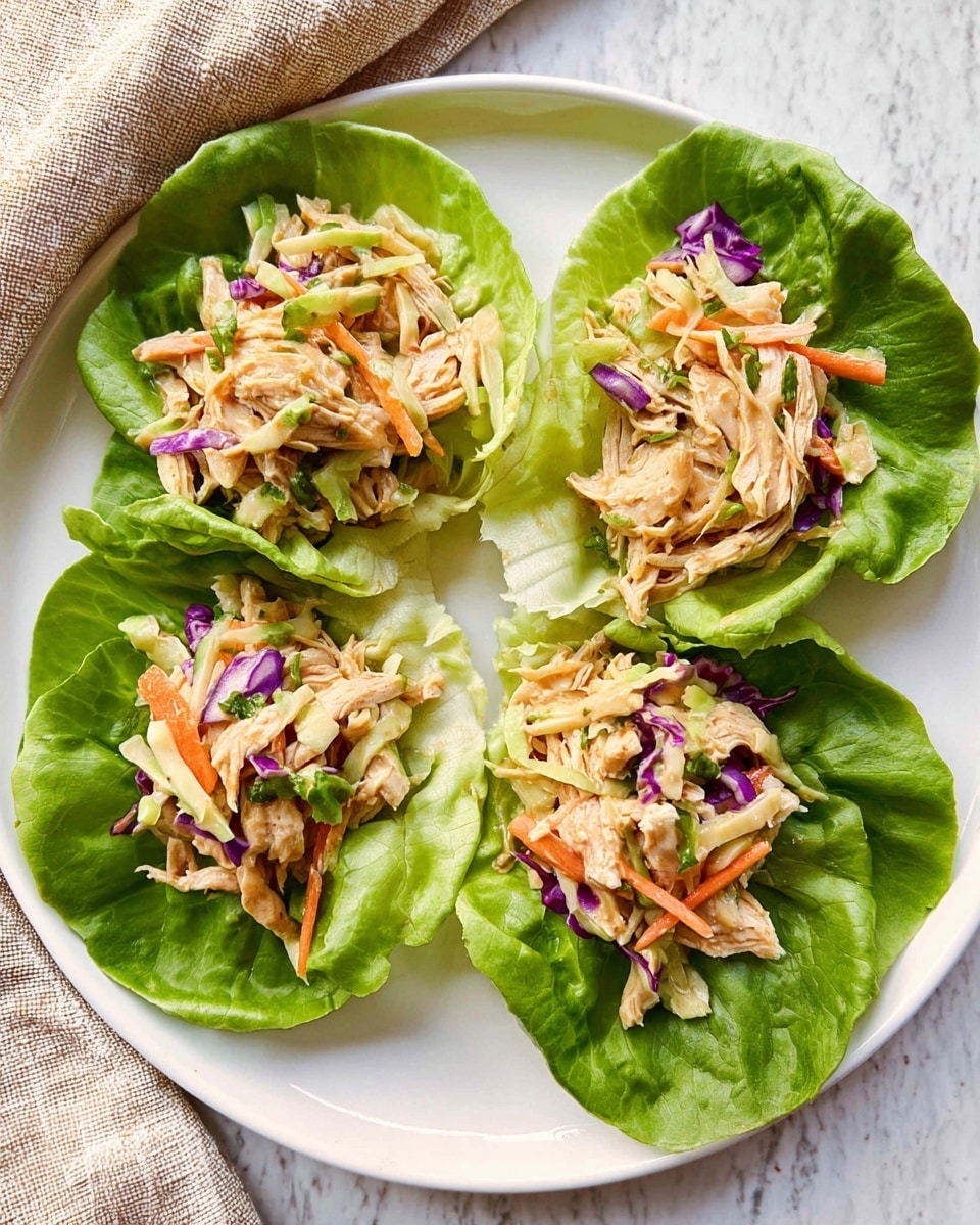 The image shows a white round plate placed on a white marbled surface, holding four green leafy lettuce wraps arranged in a circle with a space in the middle. Each lettuce leaf is fresh and bright green, forming a bowl-like shape. Inside the lettuce wraps, there are shredded light-brown cooked chicken pieces mixed with thin strips of purple and light green cabbage, orange carrot, and some small bits of green herbs. The mix looks fresh and colorful with a slight shine. In the top left corner, a beige cloth is partially visible. The photo taken with an iphone --ar 4:5 --v 7