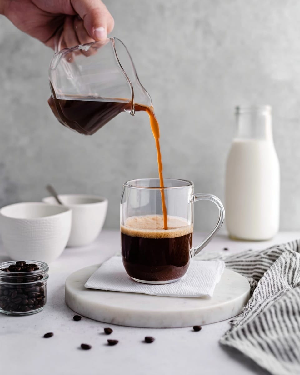 A clear glass mug with a handle is placed on a white napkin on top of a round white marble board; dark brown coffee is being poured into the mug from a small clear glass pitcher held by an arm and a hand. The coffee is in mid-pour, showing its rich brown color and slight foam at the top. Around the scene, there are scattered dark coffee beans, two white bowls stacked on the left, a small square jar filled with dark coffee beans, a tall glass bottle of milk in the background, and a folded striped cloth on the right side, all set on a white marbled surface with a soft gray textured background. Photo taken with an iphone --ar 4:5 --v 7