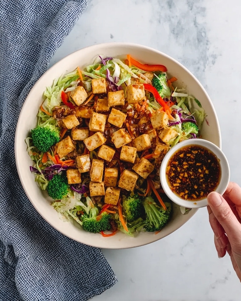 A white bowl sits on a white marbled surface, filled with a colorful salad. The bottom layer consists of fresh, shredded green and purple cabbage mixed with thin slices of orange carrot and red bell pepper strips. Bright green broccoli florets are scattered evenly over the base. On top of the vegetables lie many small, golden brown tofu cubes, slightly crispy in texture. A woman's hand is holding a small white bowl filled with dark brown sauce containing visible bits of garlic and other seasonings, pouring it over the tofu and vegetables. A blue and white checkered cloth napkin is placed next to the bowl. Photo taken with an iphone --ar 4:5 --v 7