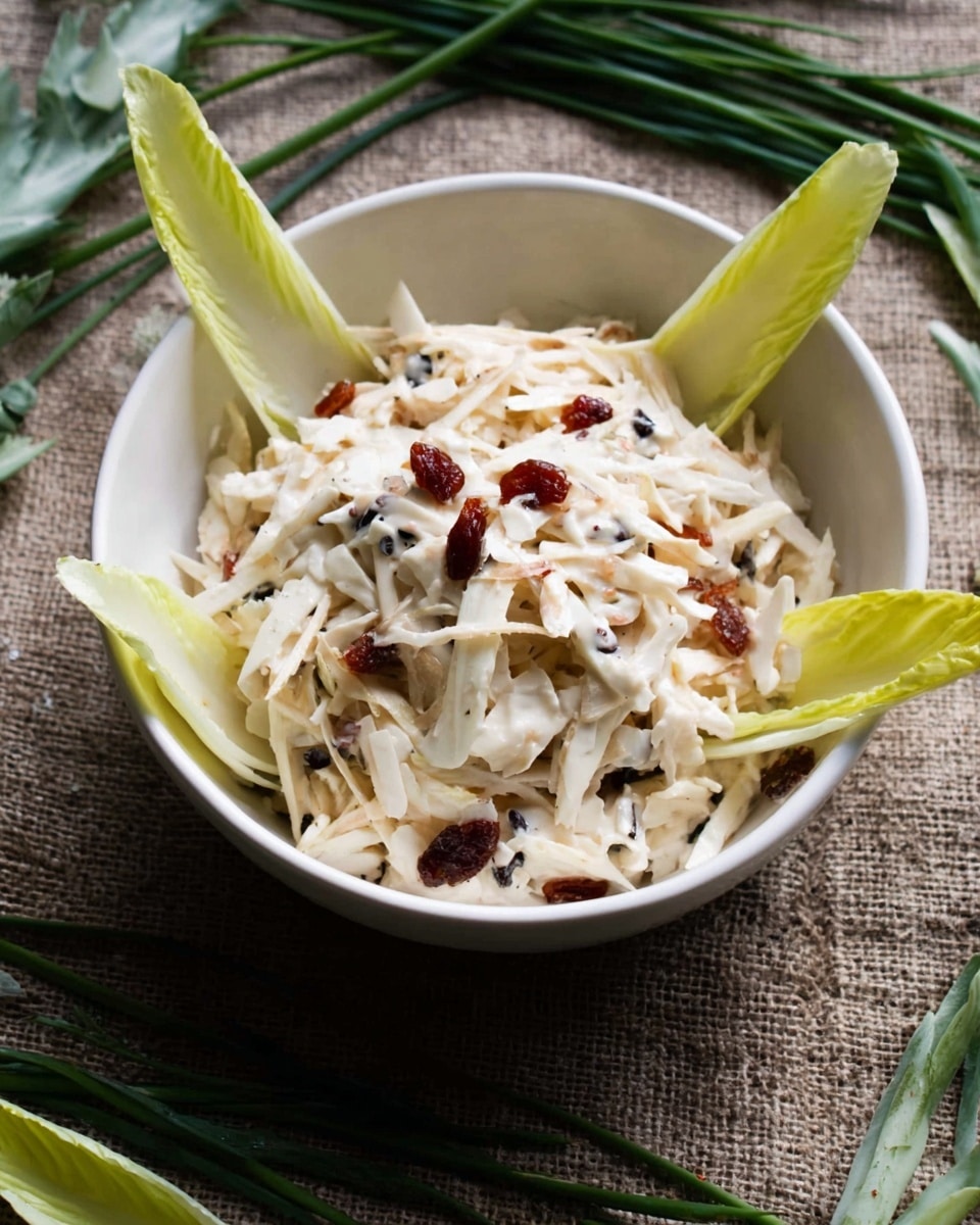 A white bowl filled with a creamy salad made of light beige shredded pieces mixed with small dark bits and red dried fruit pieces. Around the creamy salad inside the bowl, there are several light green and pale yellow endive leaves placed standing up, creating a star-like shape. The bowl sits on a rough textured brown fabric surface with green chives and some endive leaves scattered around. Photo taken with an iphone --ar 4:5 --v 7