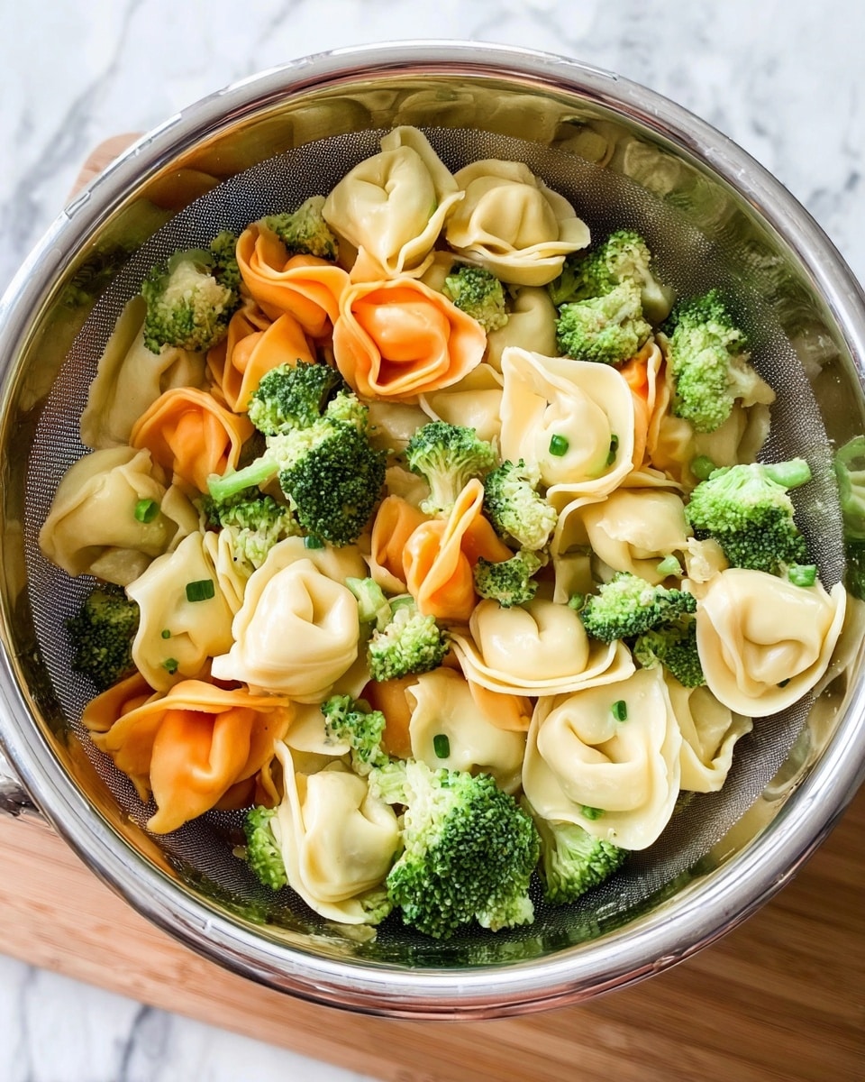 The image shows a silver metal colander filled with three colors of tortellini pasta - white, orange, and green. The tortellini are mixed with small bright green broccoli florets scattered evenly throughout. The pasta has smooth and slightly wrinkled texture with some pieces folded tightly. The colander sits on a light brown wooden surface, and the background is a white marbled texture. The photo taken with an iphone --ar 4:5 --v 7