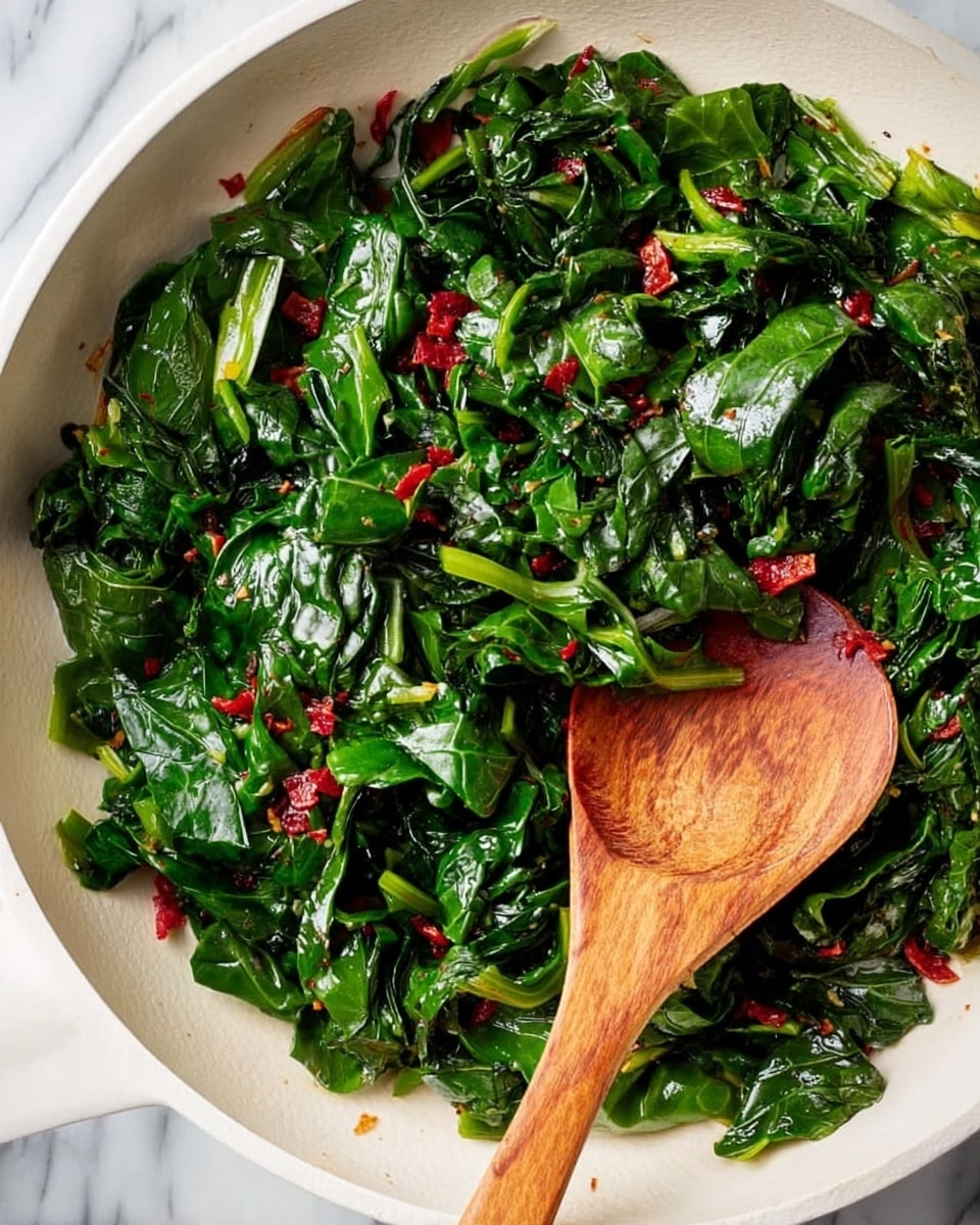 A white pan filled with bright green cooked leafy greens mixed with small red pieces, showing a fresh and shiny texture. The greens have a slightly wrinkled surface with folded layers, while the red pieces appear soft and scattered evenly among the leaves. A light-colored wooden spoon with visible grain rests inside the pan, partially covered by the greens. The background features a white marbled surface. photo taken with an iphone --ar 4:5 --v 7