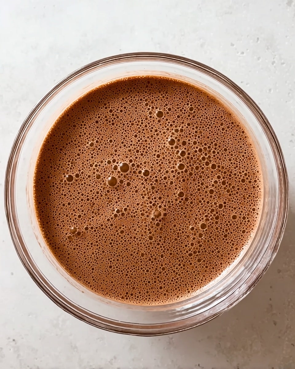 A clear glass bowl filled with a smooth layer of chocolate mixture that is light brown in color with many small bubbles spread evenly across the surface, sitting on a white marbled texture. The bowl is round and transparent, showing the uniform texture of the chocolate inside, with the rim of the bowl clearly visible. Photo taken with an iphone --ar 4:5 --v 7