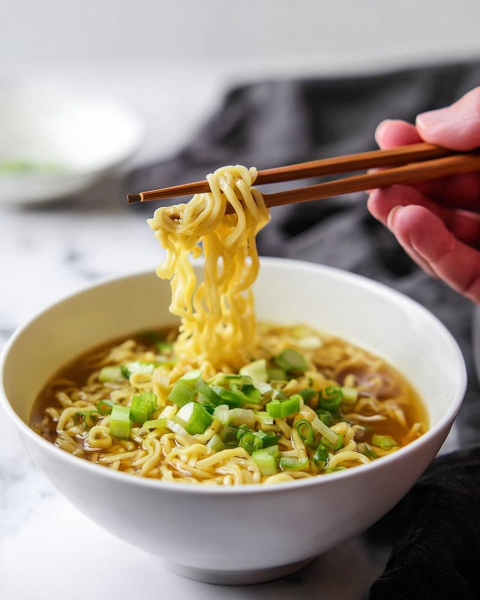 A white bowl filled with three layers: the bottom layer is light brown broth, the middle layer is yellow cooked noodles with soft texture, and the top layer is bright green chopped scallions scattered evenly. A woman's hand is holding wooden chopsticks, lifting some noodles from the bowl. The whole scene sits on a white marbled surface with a soft blurred background and a hint of another white dish in the back. photo taken with an iphone --ar 4:5 --v 7