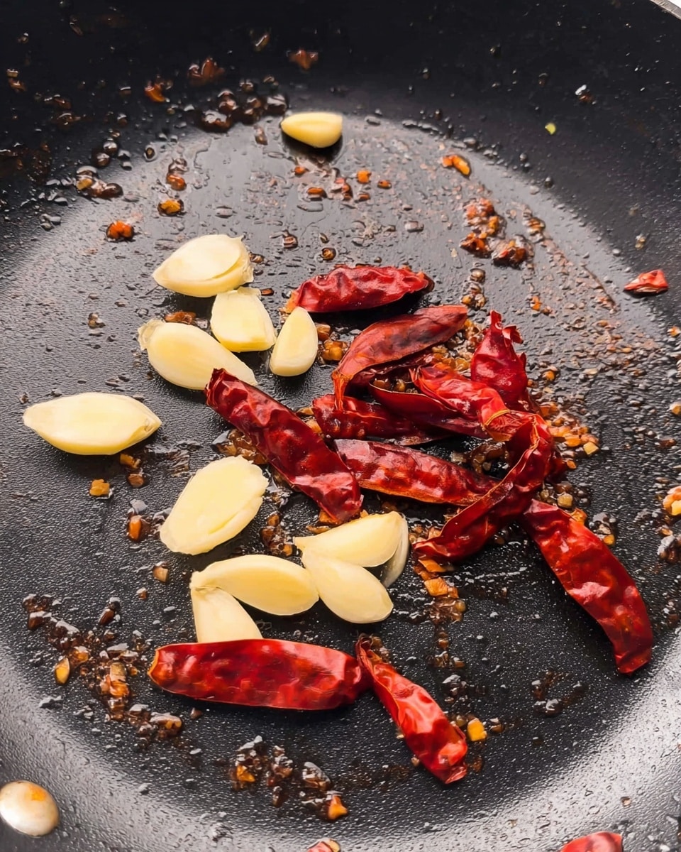 A close-up view of a black pan with pieces of sliced garlic and whole dried red chili peppers spread around the center. The garlic slices are pale yellow and smooth, while the dried chilies are bright red with a dry, wrinkled texture. Small brown oil spots and bits of cooked residue spread unevenly on the pan's surface, with a slight shine from the oil. The background shows a hint of black edges of the pan. photo taken with an iphone --ar 4:5 --v 7