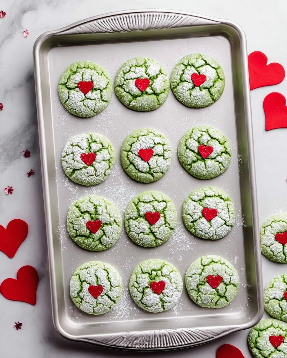 The image shows a silver baking tray with twelve round, green cookies arranged in four rows of three. Each cookie has a cracked surface dusted with white powdered sugar, giving it a textured look. In the center of each cookie, there is a small, flat red heart decoration. The tray is placed on a white marbled surface with a few more red heart decorations scattered around. Some green cookies can also be seen partially on the right side of the image. Photo taken with an iphone --ar 4:5 --v 7