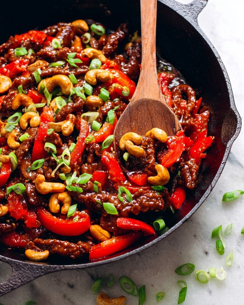 A close-up view of a black cast iron pan filled with a stir-fry dish showing 4 main layers: the base layer is browned cooked meat strips coated in a shiny dark brown sauce, the second layer is bright red sliced bell peppers that add a pop of color, the third layer includes light golden toasted cashews scattered throughout, and the top layer is fresh sliced green onions sprinkled on top, adding a fresh green contrast. A wooden spoon is partially submerged in the pan among the ingredients. The pan is placed on a white marbled surface with some green onion slices scattered around. photo taken with an iphone --ar 4:5 --v 7