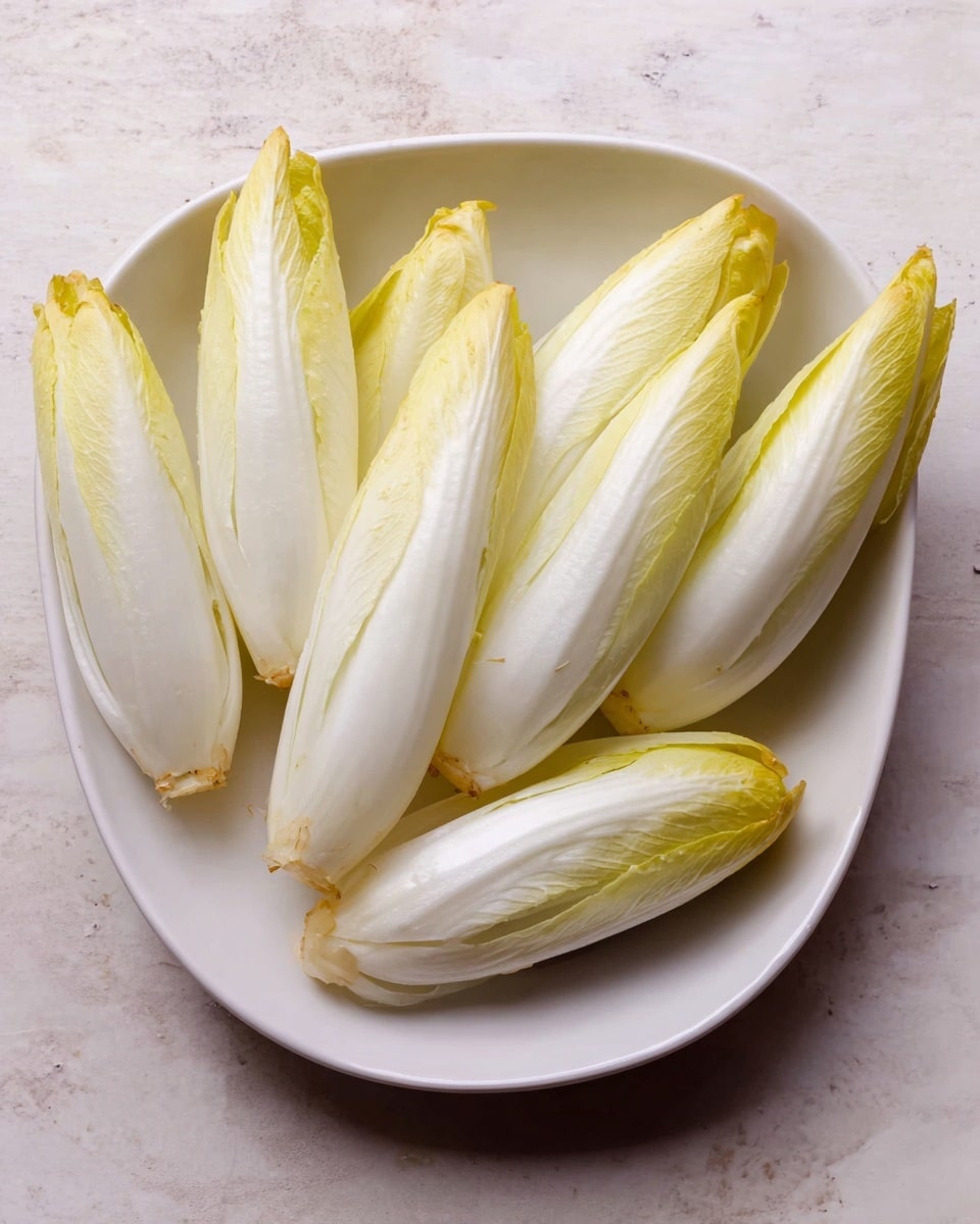 A white dish holds several whole endive leaves arranged neatly, with their pale yellow tips pointing outward and bottom ends gathered toward the center; the leaves are smooth with a slight shine, showing a gradient from creamy white at the base to light yellow near the tips, resting on a white marbled surface texture photo taken with an iphone --ar 4:5 --v 7