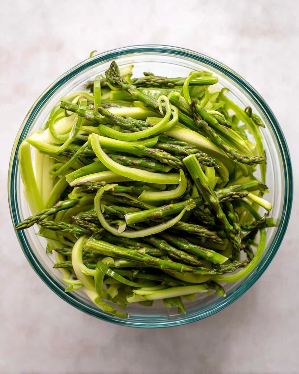 A clear glass bowl filled with many thin asparagus strips, some are curled and some straight, showing vibrant green colors with a mix of light and dark green shades. The asparagus strips are arranged loosely and fill the bowl to the top, with some tips pointing outwards. The bowl sits on a white marbled surface, giving a clean and fresh look. photo taken with an iphone --ar 4:5 --v 7
