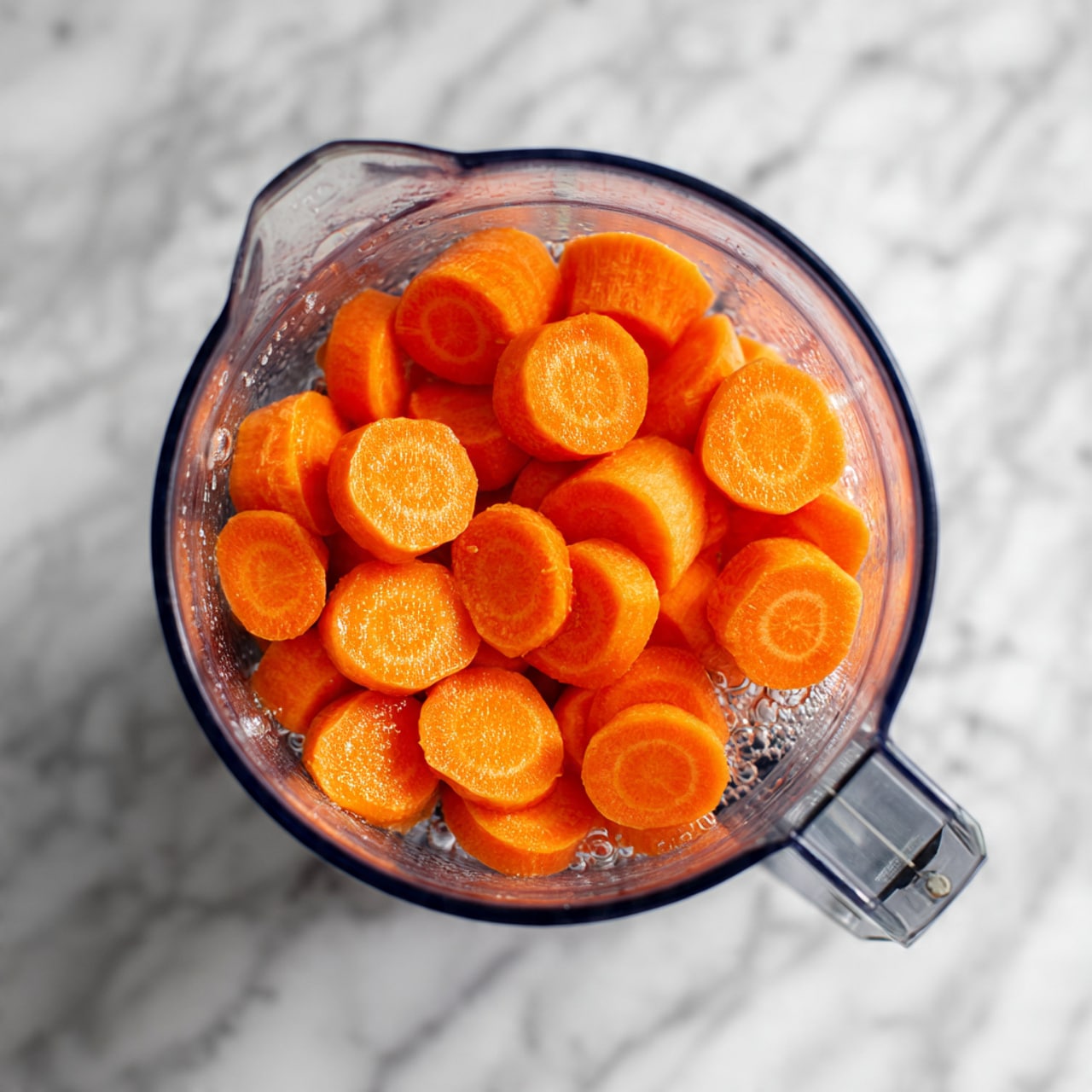 The image shows a white blender cup filled with many pieces of bright orange carrot, cut into small thick round slices, almost filling the blender. The carrot pieces have a smooth texture and are covered with clear water, with a few bubbles floating on the surface. The blender sides are clear, showing the carrots fully immersed in the water, and a metal blade is seen at the bottom right corner inside the blender. The background is a white marbled texture photo taken with an iphone --ar 4:5 --v 7