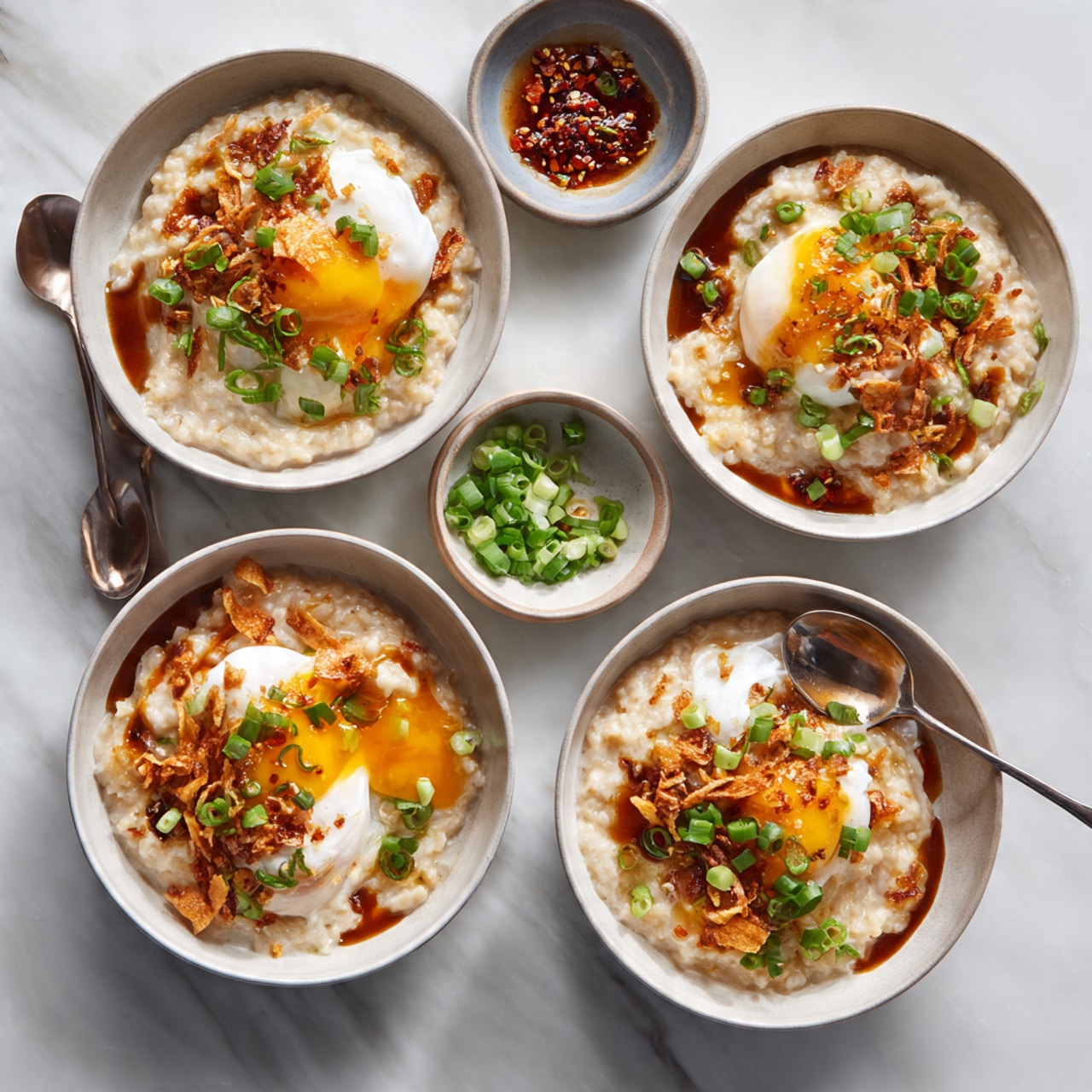 The image shows four white bowls filled with a layered dish placed on a white marbled surface. Each bowl contains a base of creamy oatmeal or porridge with a light beige color and soft, slightly chunky texture. On top of the oatmeal is one poached egg per bowl, with glossy white and slightly runny bright orange yolk spreading lightly in some bowls. The eggs are sprinkled with chopped green onions and crispy golden fried onions, adding a mix of green and dark brown textures and colors. A dark savory sauce is drizzled over the eggs, pooling around edges in some bowls. Small bowls with additional chopped green onions and chili flakes, and spoons are placed near the four bowls. The photo is taken with an iphone --ar 4:5 --v 7