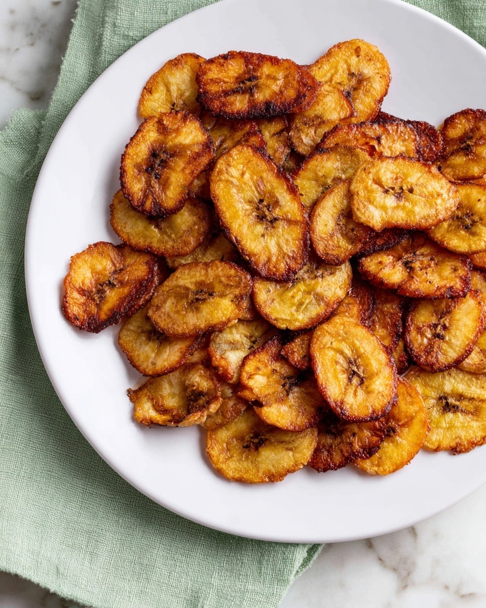 A white plate holds many slices of fried plantains. Each slice is oval-shaped with a golden brown color and darker crispy edges, showing a slightly rough and crunchy texture. Some slices have small darker brown spots scattered, giving a varied toasted look. The plate is set on a white marbled surface with a soft green cloth partially visible under the plate. The fried plantains are evenly spread, filling most of the plate, with a rustic and homemade appearance. photo taken with an iphone --ar 4:5 --v 7
