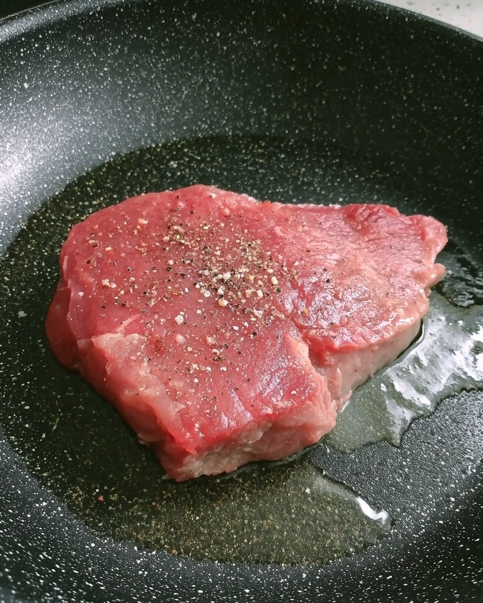 A close-up view of a single raw steak piece in a frying pan, the steak is pink with some light marbling and sprinkled lightly with salt and pepper. The pan is black with white speckles, and there is oil pooling around the steak, making its surface shiny. The background is a white marbled texture. photo taken with an iphone --ar 4:5 --v 7