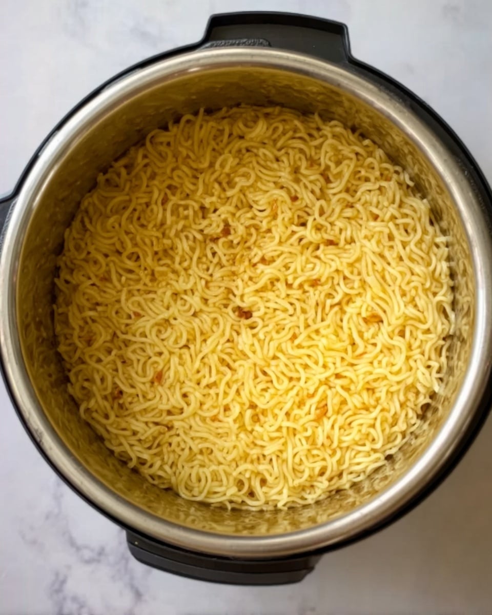 A metal pot filled with a single layer of cooked yellow noodles evenly spread out, with some small browned bits visible throughout the noodles. The pot is placed on a white marbled surface, and a black part of the appliance is slightly visible on the top edge. The noodles have a smooth texture with light shine, filling the pot completely. Photo taken with an iphone --ar 4:5 --v 7