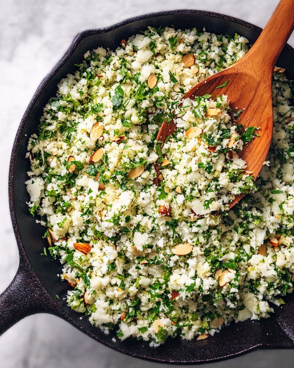 A black cast iron pan filled with a mixture that looks like finely chopped white cauliflower with bright green herbs and small light brown almond slices mixed throughout. A wooden spoon with a round handle is scooping some of the mixture, showing the soft, crumbly texture of the cauliflower and herbs blend. The background surface is a white marbled texture. photo taken with an iphone --ar 4:5 --v 7