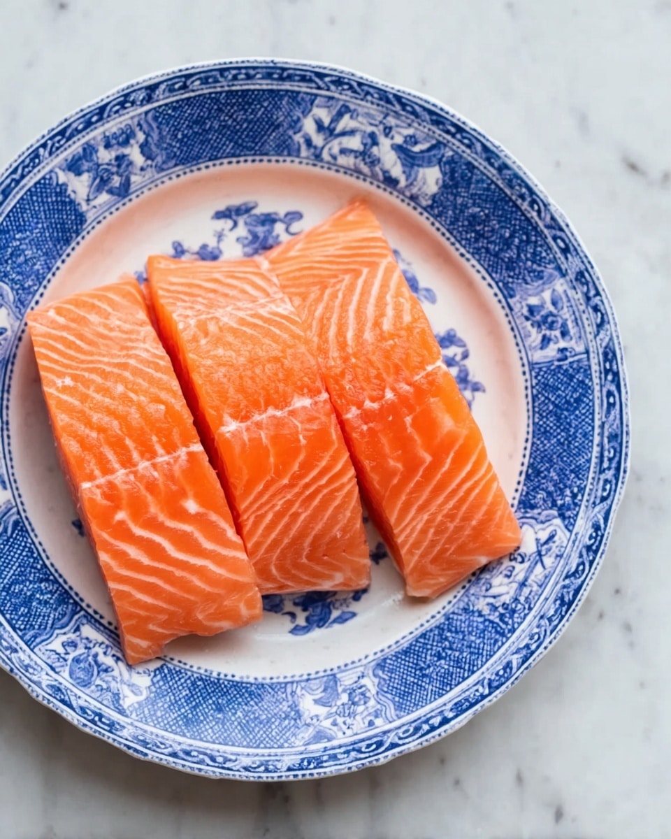 Three thick, fresh orange salmon fillets lay side by side in a white plate with blue traditional patterns around its edge, showing smooth and slightly marbled textures with faint white lines running through the fish. The plate rests on a white marbled surface that looks clean and soft. photo taken with an iphone --ar 4:5 --v 7