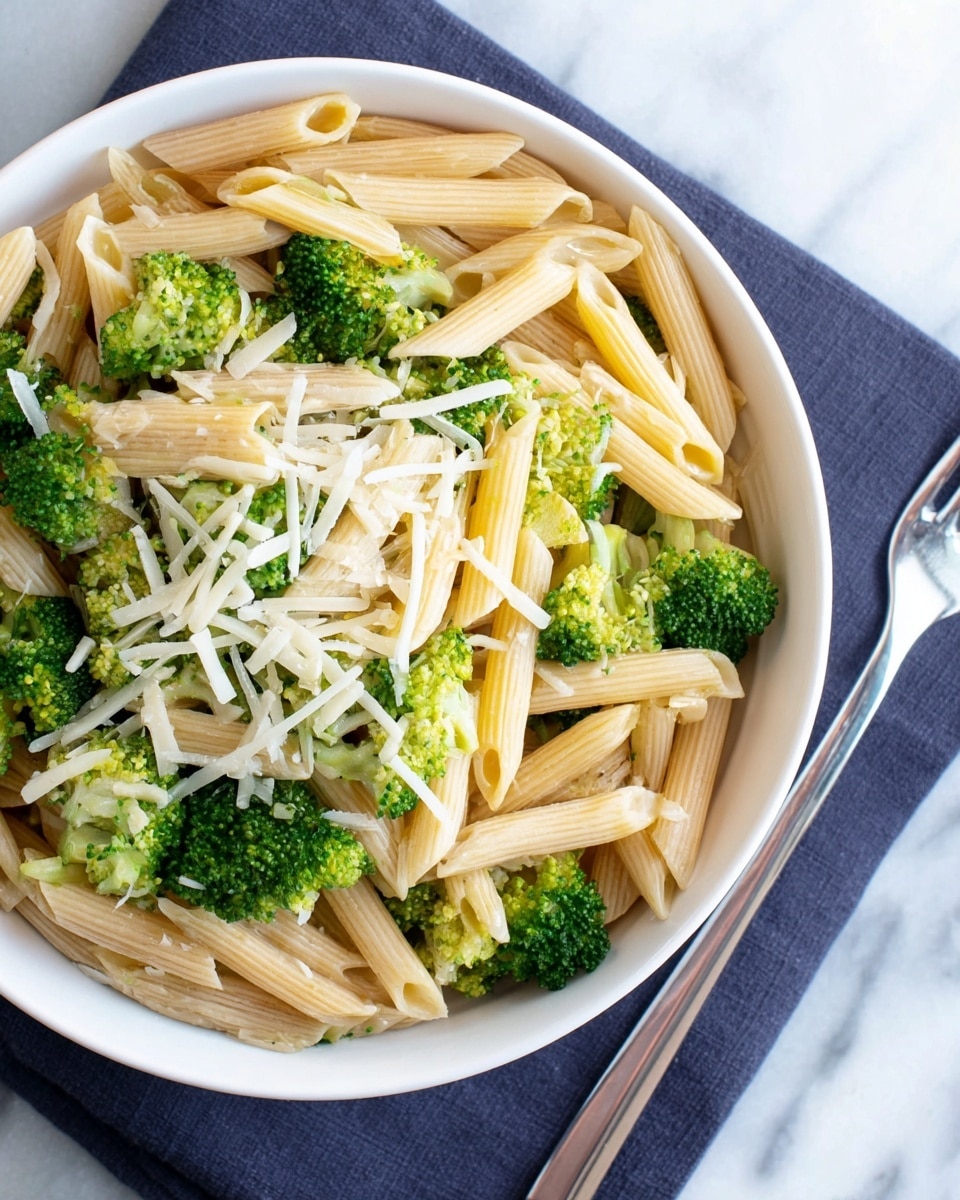 A white bowl is filled with three layers of food: the bottom layer is light beige penne pasta, the middle layer is bright green broccoli florets scattered evenly, and the top layer has thin white shavings of cheese spread across. The pasta looks soft and plain, and the broccoli pieces are small and fresh. The bowl sits on a dark blue cloth on a white marbled surface, with a silver fork visible on the right side. photo taken with an iphone --ar 4:5 --v 7