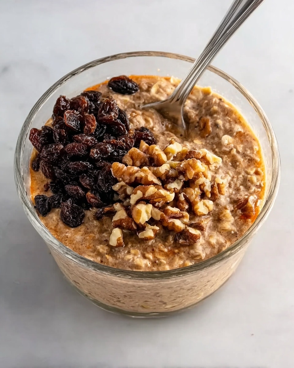A clear glass bowl filled with a thick oatmeal mixture that is light brown with visible small chunks of oats and nuts, topped with a pile of dark raisins on the left and a smaller pile of chopped walnuts on the right. A silver spoon is resting inside the bowl near the top edge, angled toward the right side. The bowl is placed on a white marbled surface photo taken with an iphone --ar 4:5 --v 7