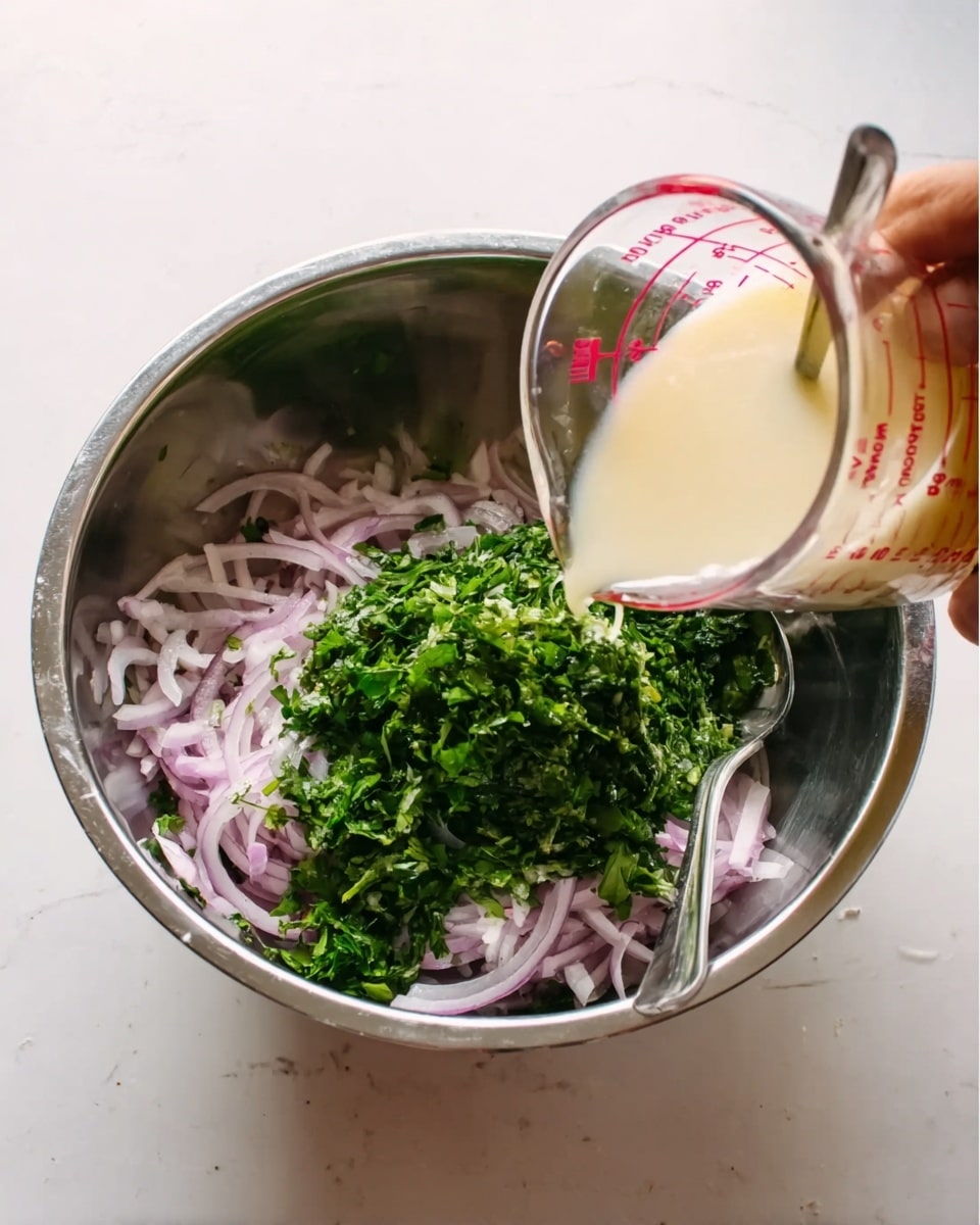 A stainless steel bowl sits on a white marbled surface, filled with layers of thinly sliced pale purple onions and a pile of finely chopped bright green herbs in the center. Above the bowl, a woman's hand is pouring a light yellow creamy liquid from a clear measuring cup with red markings over the onions and herbs. A silver spoon rests inside the bowl, partially submerged in the onion layer. The scene is brightly lit and clean, photo taken with an iphone --ar 4:5 --v 7