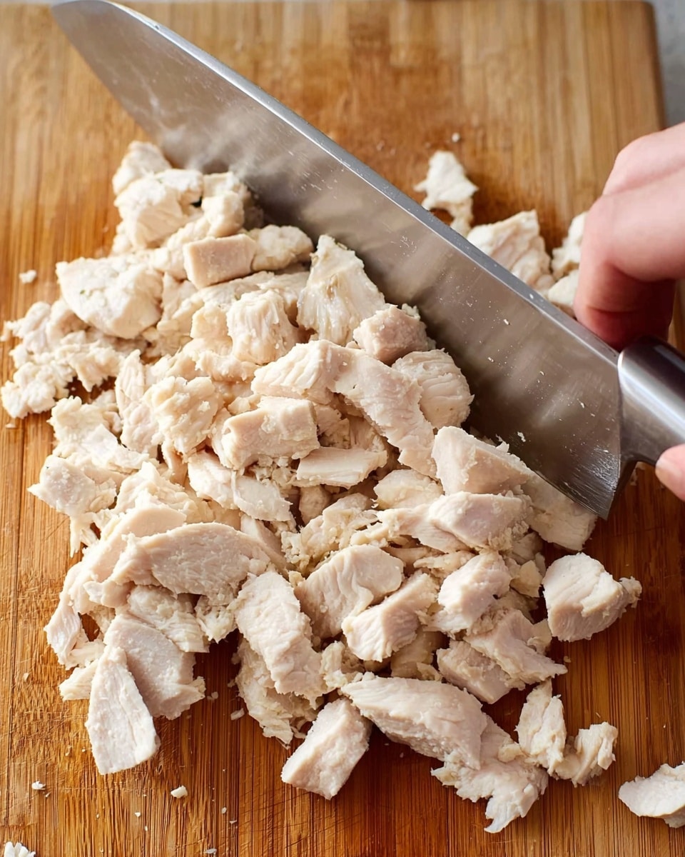 The image shows a close-up of cooked white chicken pieces being chopped on a wooden cutting board. The chicken is broken into small to medium rough chunks, filling most of the board. A large silver knife cuts through the chicken horizontally. A woman's hand grips the knife handle firmly on the right side of the frame. The wooden board has a natural light brown color with some visible wood grain texture. The photo focuses on the texture and size of the chopped chicken pieces. photo taken with an iphone --ar 4:5 --v 7