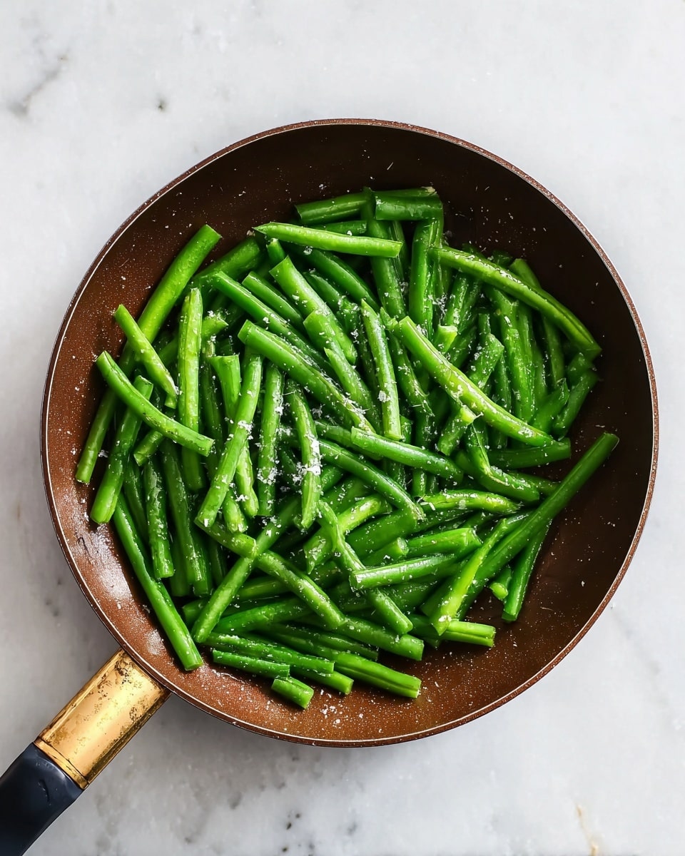 A round brown pan filled with a single layer of bright green beans that have been sliced in half, some sprinkled lightly with coarse salt. The pan has a slightly worn handle with a mix of gold and black colors. The background is a white marbled surface, giving a clean and fresh look. photo taken with an iphone --ar 4:5 --v 7