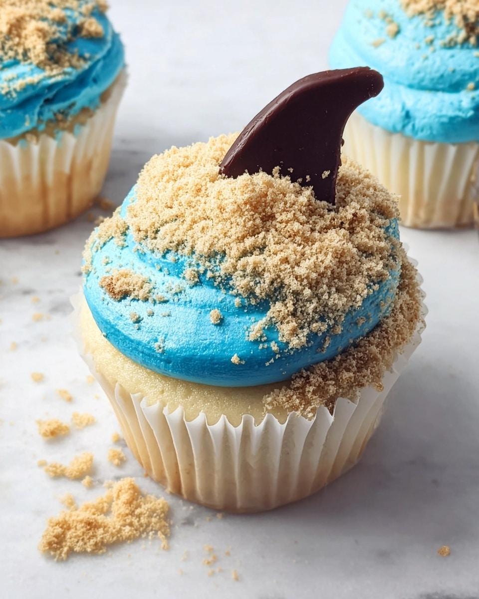 A vanilla cupcake in a white paper wrapper sits on a white marbled surface. The cupcake has one main layer of light golden cake topped with a thick layer of bright blue frosting. Most of the frosting is covered by a rough layer of light brown crumbs, with a few crumbs scattered around it on the surface. A dark brown, curved chocolate piece stands upright in the frosting near the back. Another cupcake with the same blue frosting but no crumbs is partially visible to the right. Photo taken with an iphone --ar 4:5 --v 7