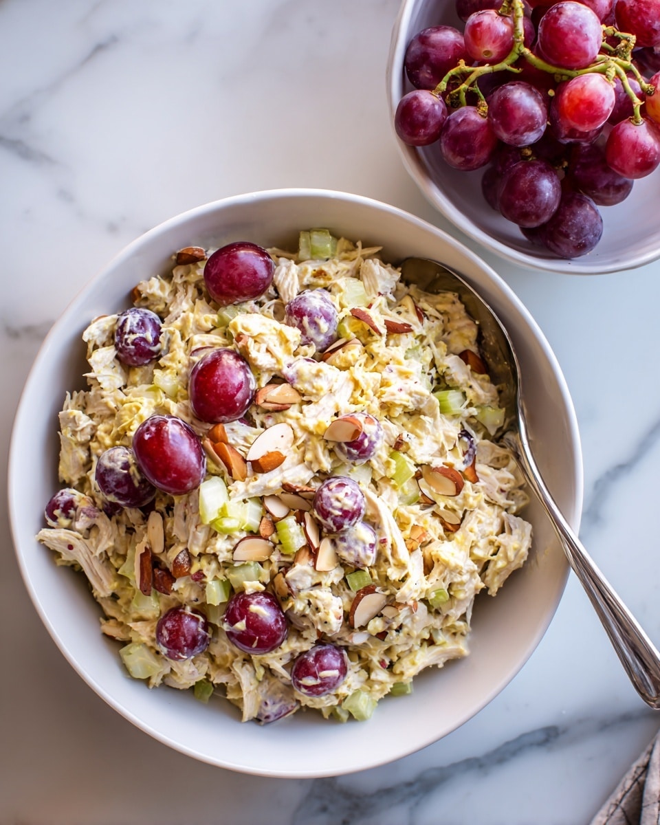 A large white bowl filled with a creamy chicken salad that has three main layers: the base is shredded chicken in a light yellow dressing, mixed evenly with small pieces of crunchy celery that appear light green; scattered on top are halved red grapes adding shiny, smooth red and purple pops; the surface shows slices of pale brown almonds for texture. The bowl sits on a white marbled surface with a silver spoon inside. Next to it, a smaller white bowl holds a bunch of red grapes. Photo taken with an iphone --ar 4:5 --v 7