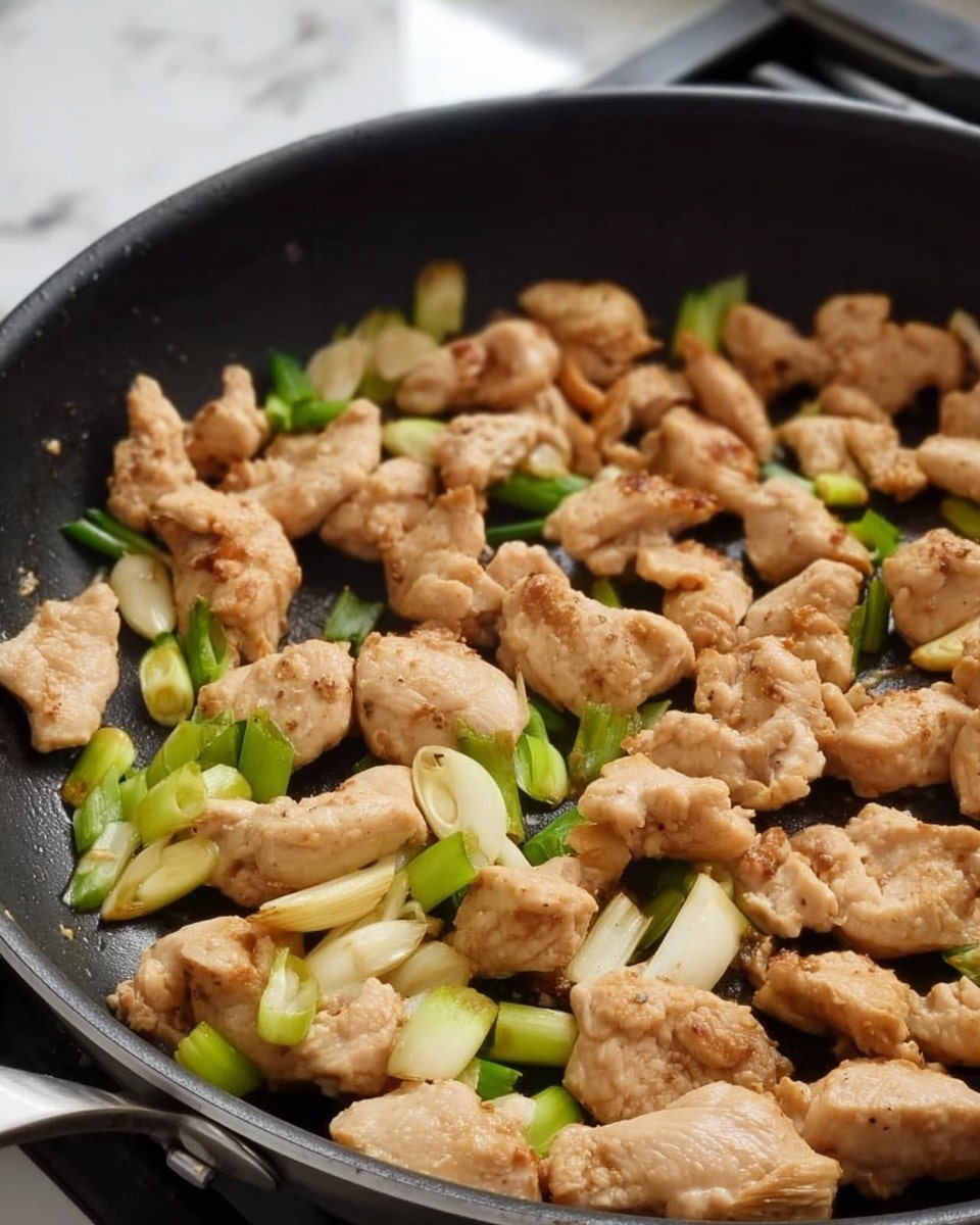 A close-up view of a black frying pan filled with multiple small pieces of light brown cooked chicken spread evenly across the pan. The chicken is lightly browned and mixed with slices of pale beige garlic and pieces of green onions with white and green parts. The pan is on a stove, and the background surface is white marbled texture. Photo taken with an iphone --ar 4:5 --v 7