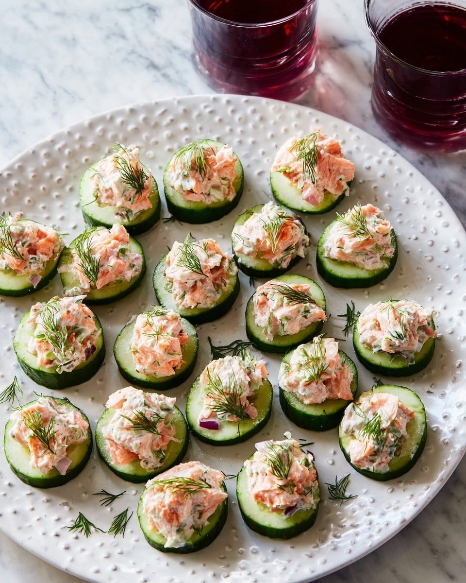 A white round plate with raised lines and dots holds many cucumber rounds topped with a creamy mixture of pinkish salmon pieces and white cream, garnished with small green dill sprigs. Each cucumber slice is thick, dark green on the outside with a pale green inside, and arranged in a scattered pattern. The creamy topping shows bits of red onion and herbs, adding texture and color. Two glasses with dark red liquid are partially visible in the upper left corner on a white marbled surface. Photo taken with an iphone --ar 4:5 --v 7