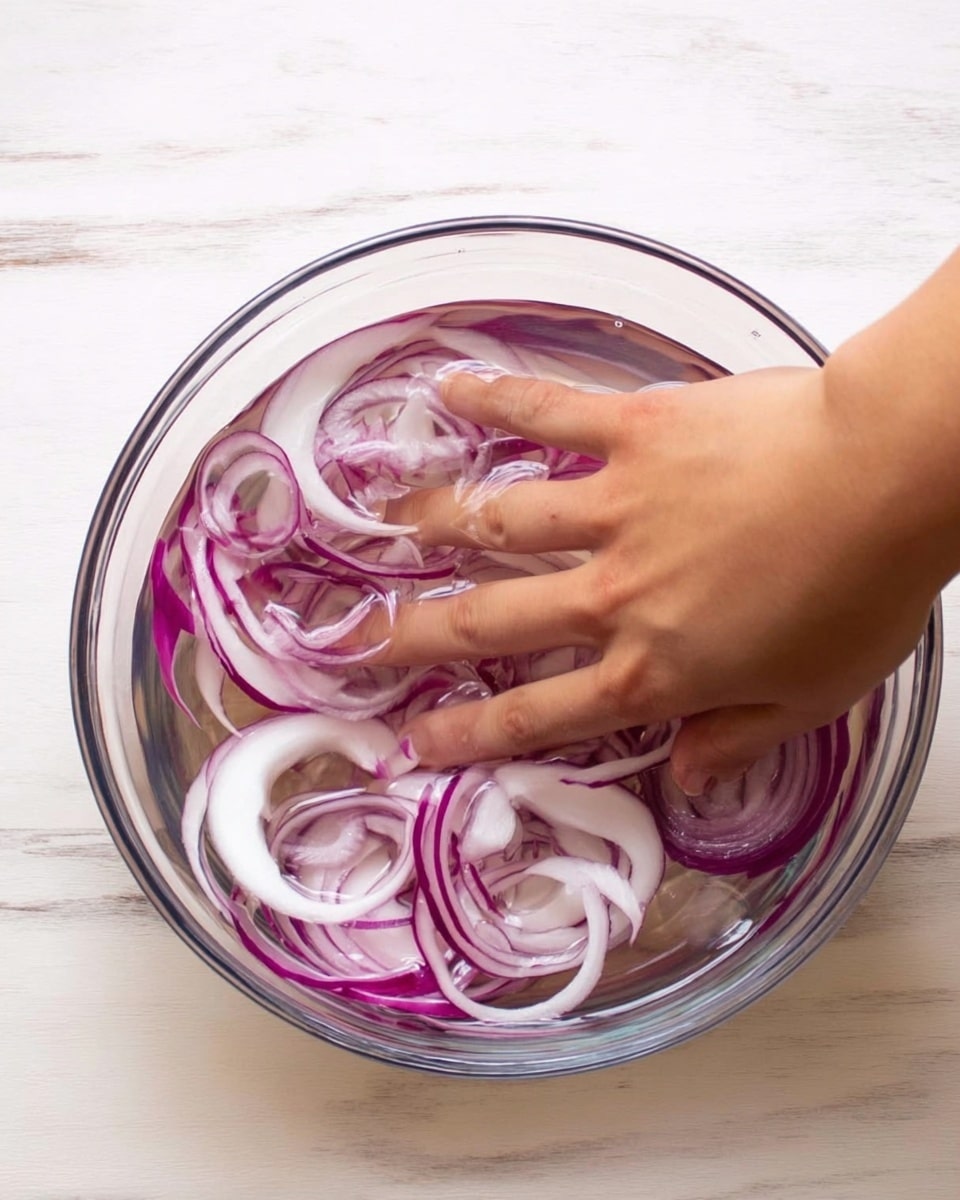 A clear glass bowl filled with water and thin slices of red onion soaking inside. A woman's hand is partially submerged in the bowl, fingers spread apart and touching the onion slices. The onion rings are white with purple edges, floating around the fingers and resting at the bottom of the bowl. The bowl is placed on a white marbled surface with soft natural lighting. photo taken with an iphone --ar 4:5 --v 7