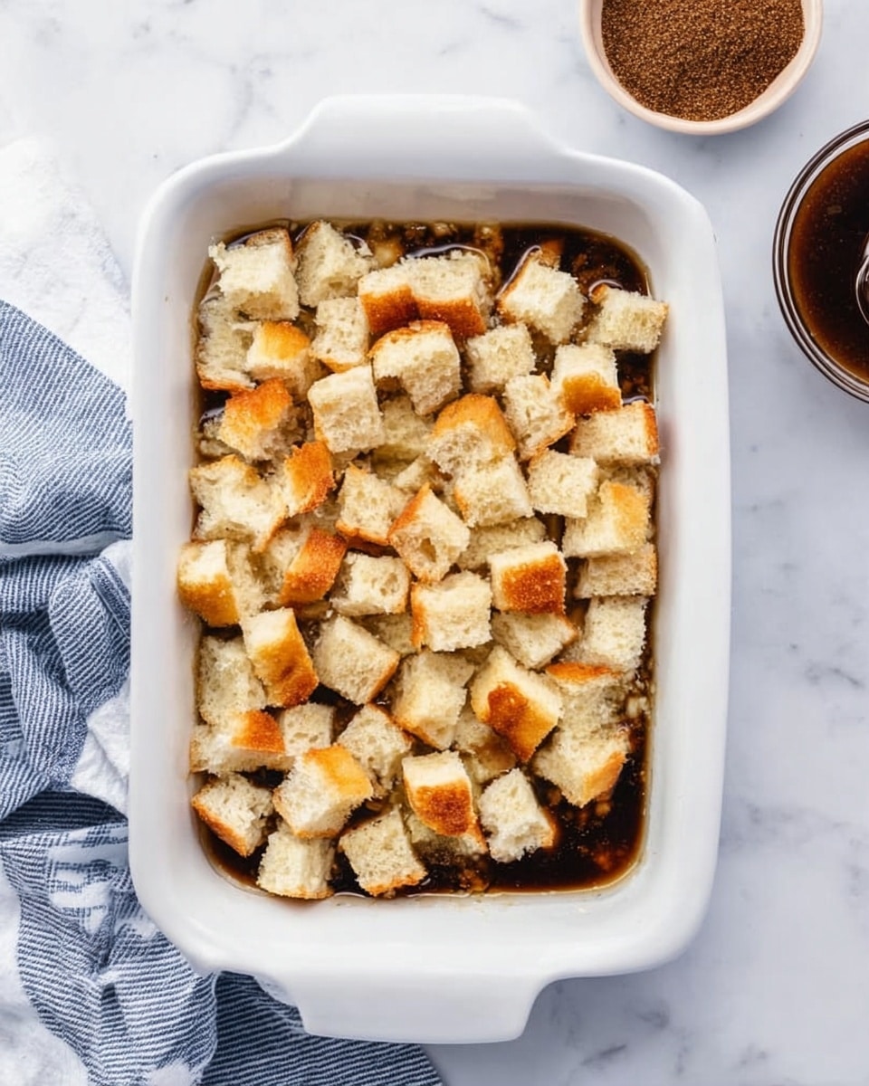 A white rectangular dish filled with one layer of small cubes of white bread with golden crusts, scattered unevenly on top of a dark brown liquid. The dish is on a white marbled surface with a blue and white striped cloth partially visible on the left side and a round container with a dark brown powder to the upper right. Photo taken with an iphone --ar 4:5 --v 7