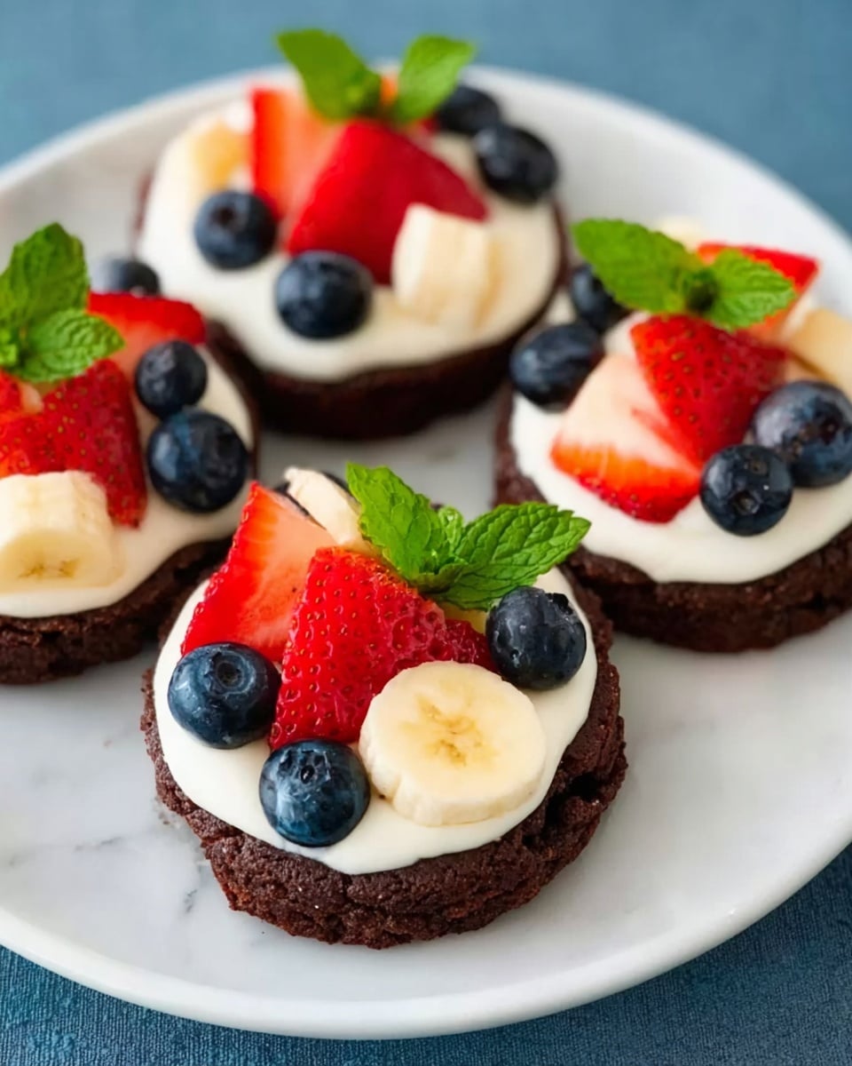 Three dark brown cookie bases are topped with a thick layer of smooth white cream, arranged on a white plate. On top of each cream layer, there are pieces of bright red strawberries, deep blue blueberries, and pale yellow banana slices scattered evenly. Each cookie is garnished with a small fresh green mint leaf, adding a touch of color and freshness. The plate rests on a white marbled surface, enhancing the bright colors of the fruit and cream. Photo taken with an iphone --ar 4:5 --v 7