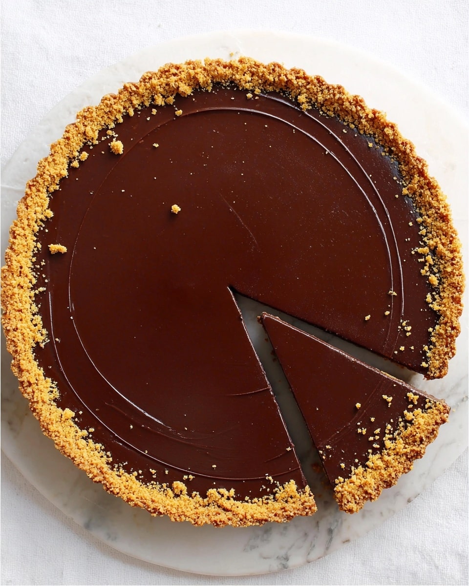 A round chocolate tart is shown from above on a white marbled surface, with a smooth, shiny, dark brown chocolate layer spread evenly over the top. A thin ring of golden brown crumb topping decorates the edge of the tart. One slice is taken out and placed slightly to the right, showing one thin, even layer of dark chocolate filling. The slice also has the same crumb topping on its edge. The overall color is rich dark brown with the contrast of the golden crumbs. Photo taken with an iphone --ar 4:5 --v 7
